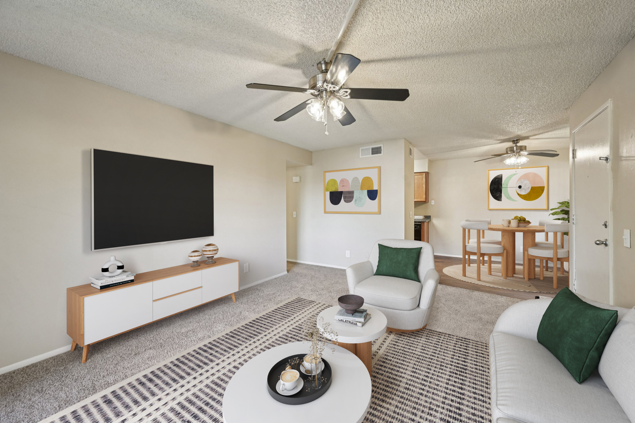 A spacious living room featuring light beige walls, a ceiling fan, and plush carpeting. The room includes a large flat-screen TV mounted on the wall, a white coffee table with decor, and a cozy sofa with green accents. A dining area with a wooden table and colorful wall art is visible in the background.