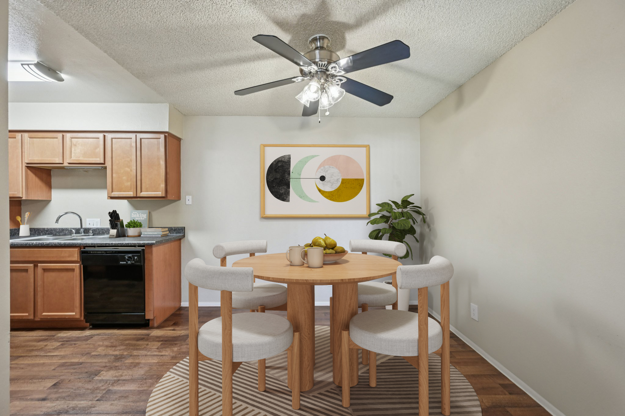A modern dining area featuring a round wooden table with four light-colored chairs. The table is adorned with a bowl of fruit. In the background, there's a kitchen with wooden cabinets and a black dishwasher. A ceiling fan hangs above, and a large abstract art piece decorates the wall next to a potted plant.