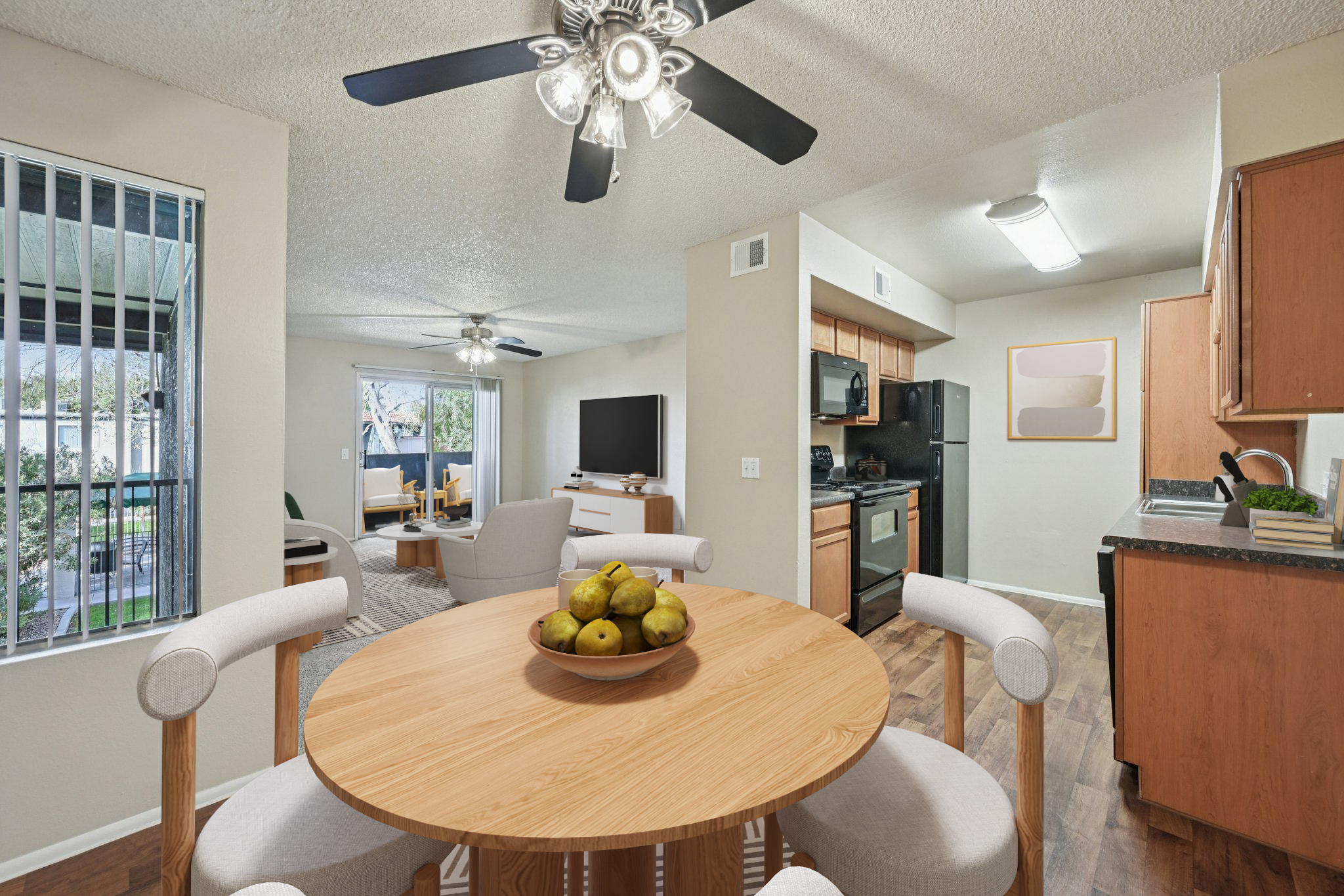 A modern dining area featuring a round wooden table with a bowl of pears at the center. The space has light-colored walls and a ceiling fan with a light fixture. In the background, a kitchen can be seen with dark appliances, and a living room area with a TV and seating is visible through an open doorway.