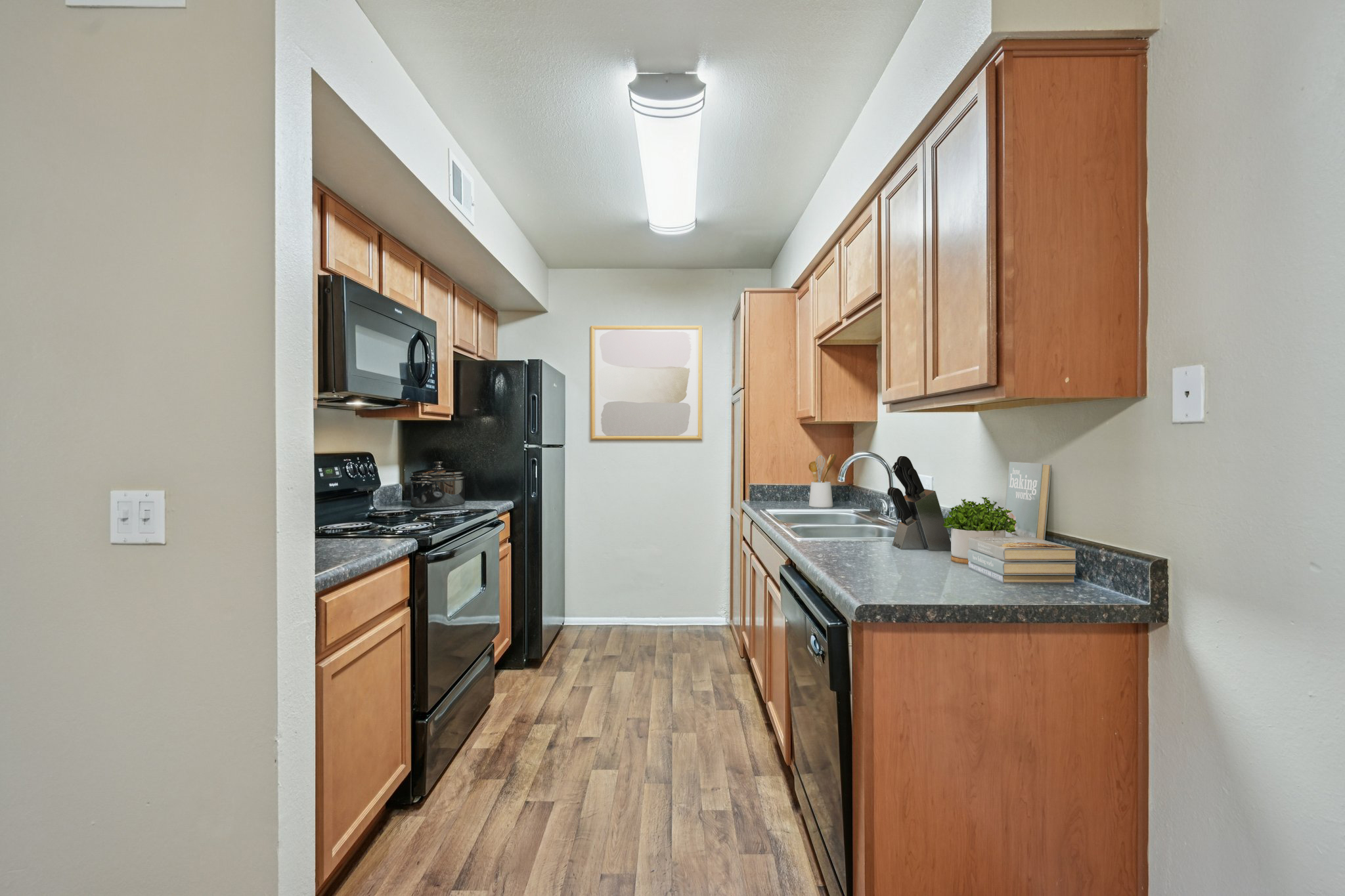 A contemporary kitchen featuring wooden cabinets, black appliances, and dark countertops. A window allows natural light to enter, illuminating the space with a modern feel. The floor is made of light wood, creating a warm atmosphere, while a small plant adds a touch of greenery.