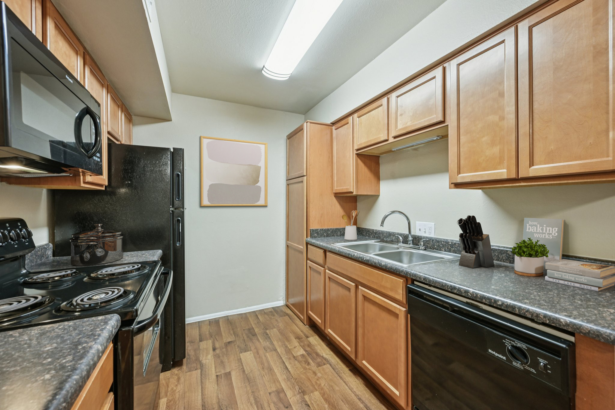 Modern kitchen featuring wooden cabinets, a black refrigerator, a black stove, and a double sink. Light-colored walls and a decorative art piece enhance the space. The countertops are dark granite, and there are kitchen utensils and a potted plant for decoration.