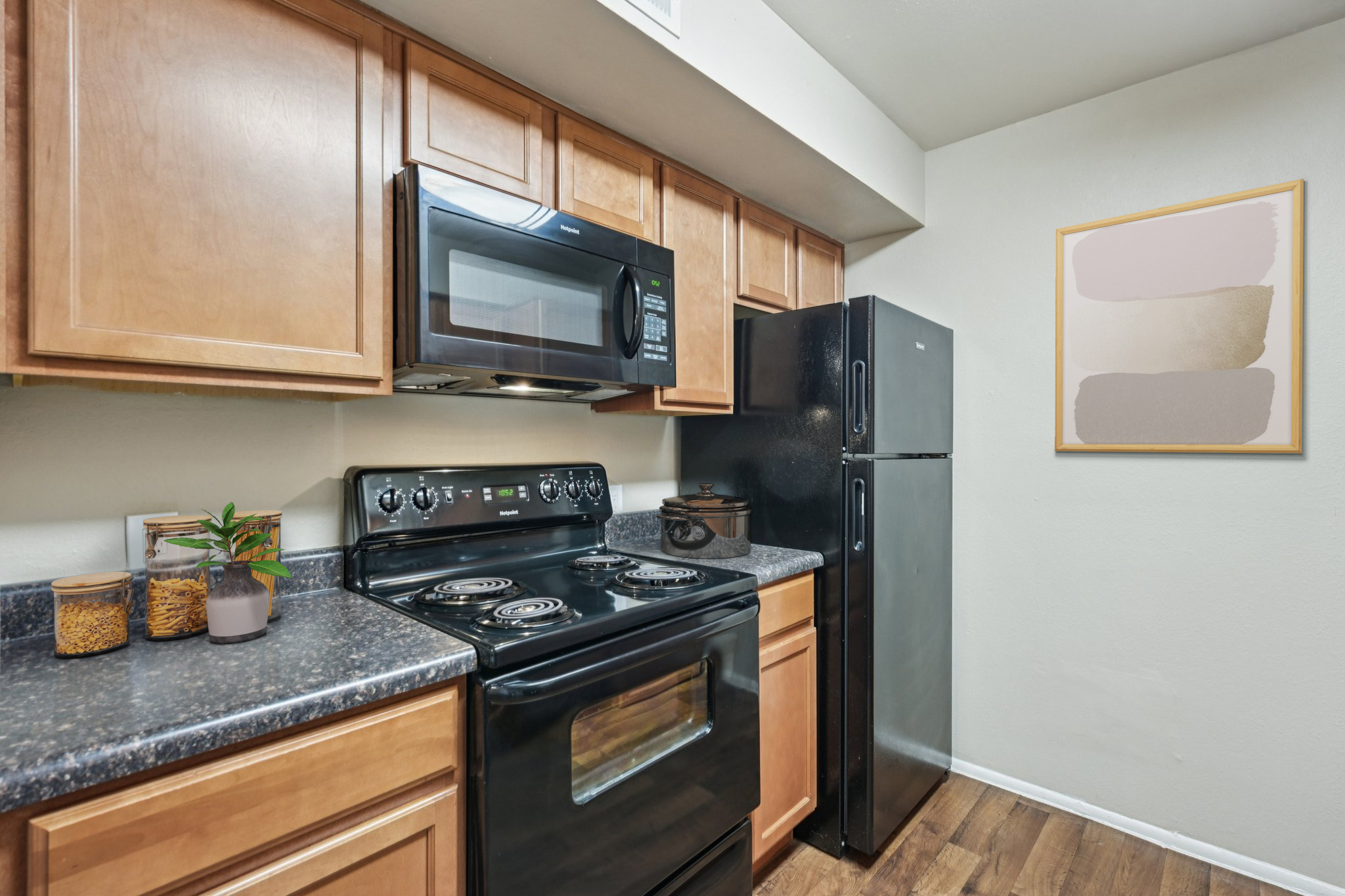 A modern kitchen featuring wooden cabinets, a black stove and microwave, and a black refrigerator. The countertops are dark, and there's a small plant and a jar of items on the counter. A minimalist framed artwork hangs on the wall, adding a decorative touch to the space.
