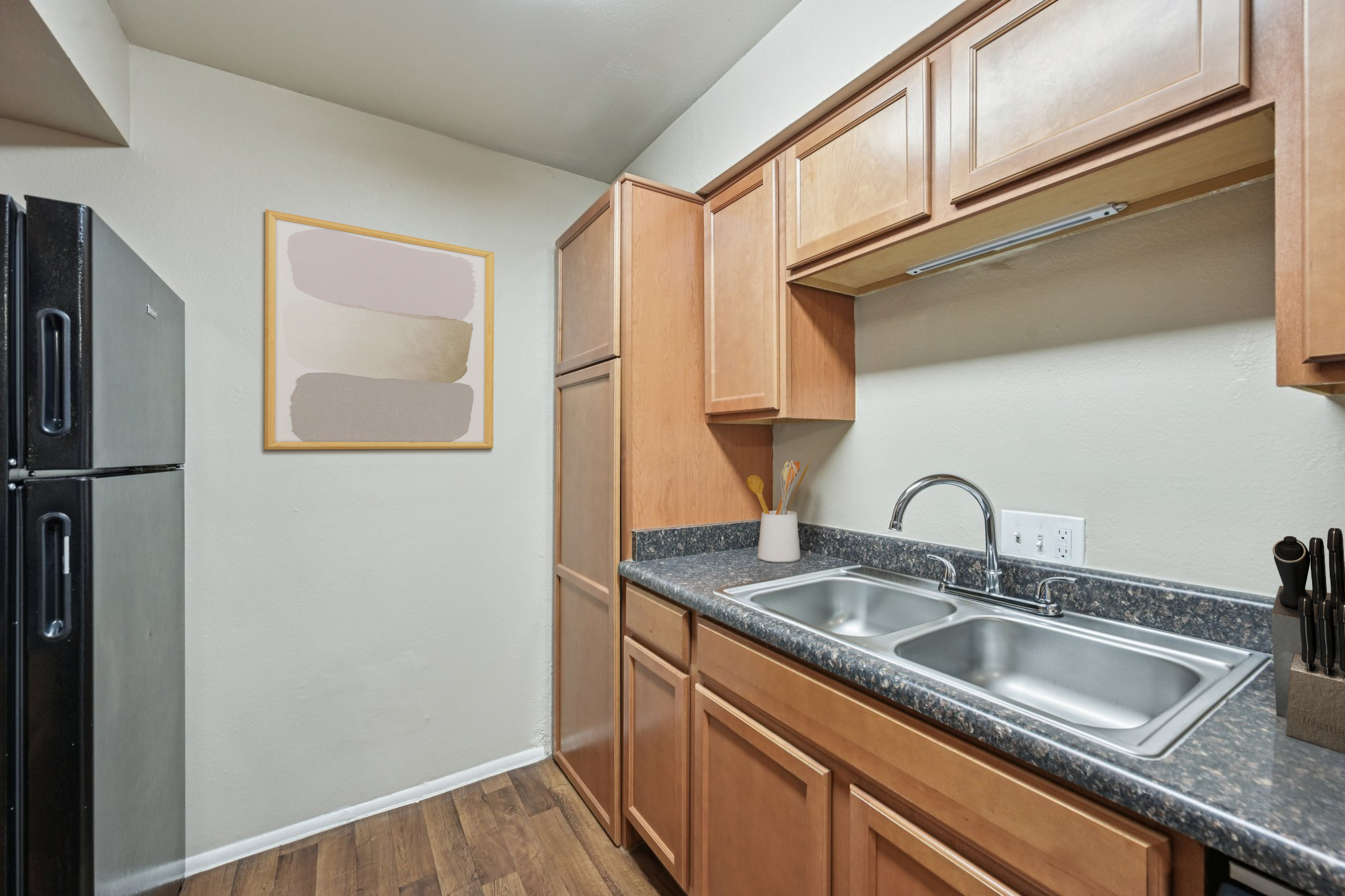 A modern kitchen featuring wooden cabinets, a stainless steel refrigerator, and a double sink. The countertop is dark with a textured surface. A piece of abstract wall art in soft colors hangs above the sink, and kitchen utensils are organized in a holder on the counter. The flooring is a warm wood finish.