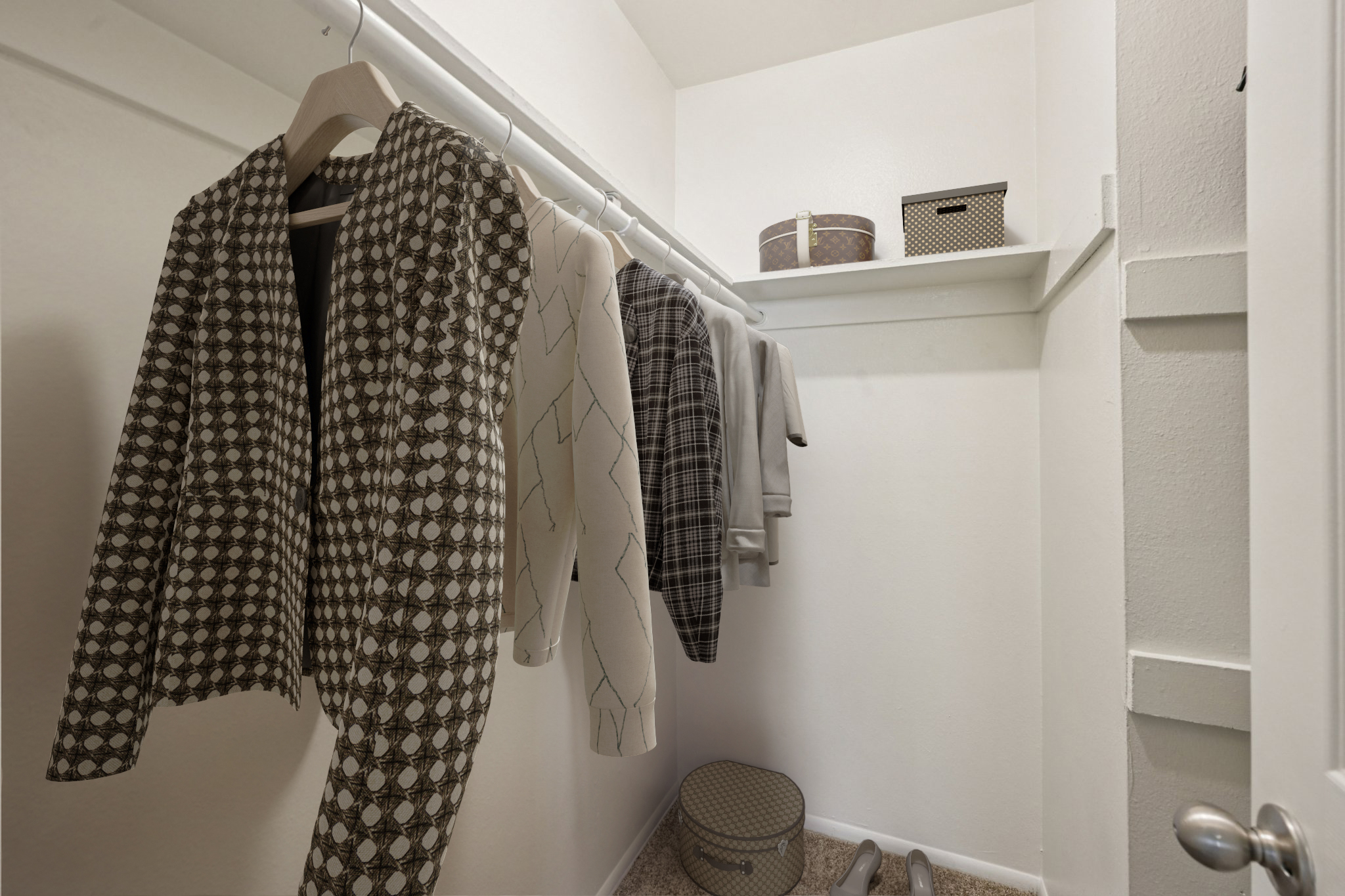 A small closet featuring a hanging rack with several patterned and solid-colored garments, including coats and sweaters. There's a shelf above with a decorative storage box and a circular storage container on the floor, accented by beige carpet.