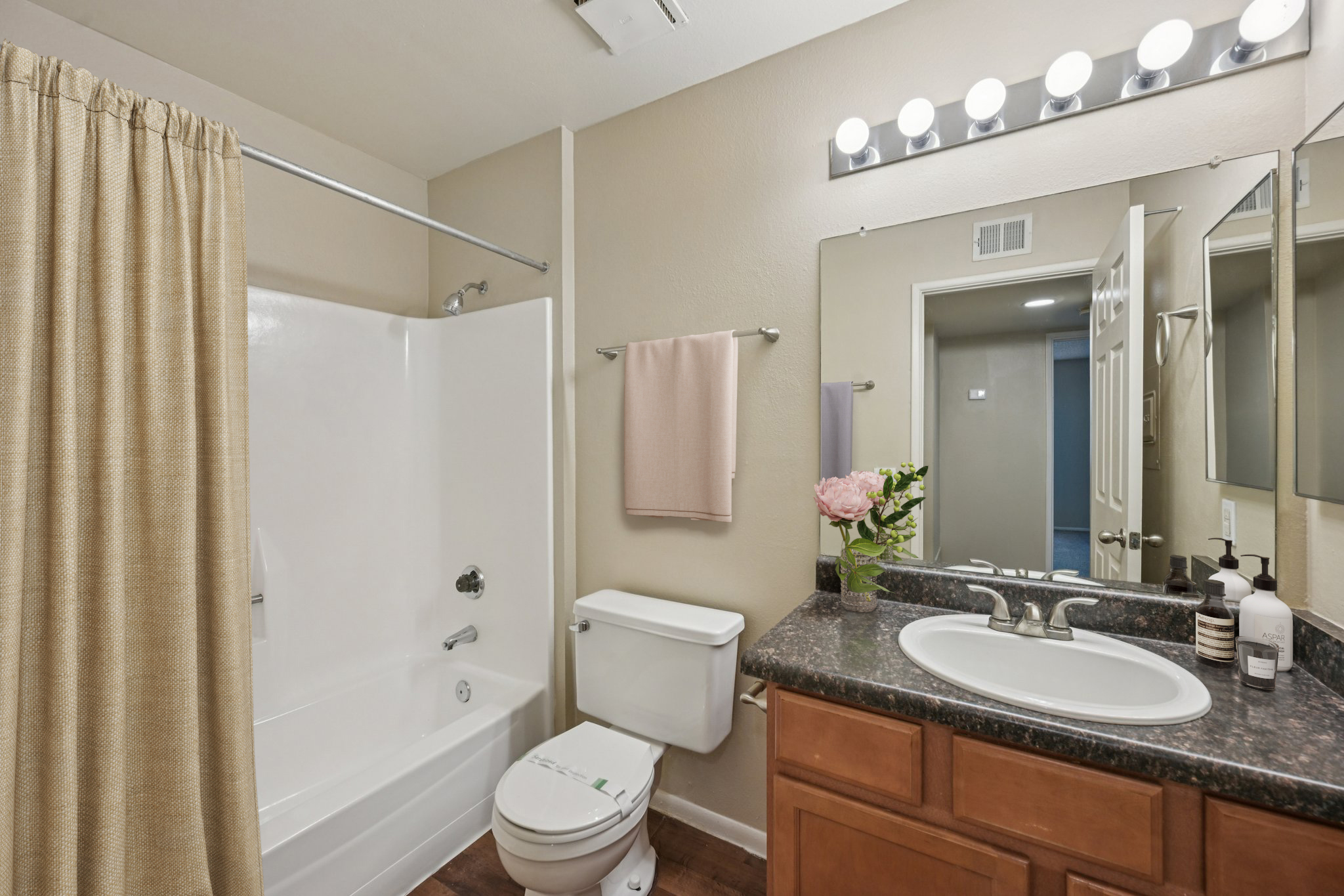 A clean and modern bathroom featuring a bathtub with a shower, a toilet, a sink with a countertop, and a small plant. The walls are painted in light tones, and there is a large mirror above the sink. Soft lighting is provided by a row of light fixtures above the mirror.