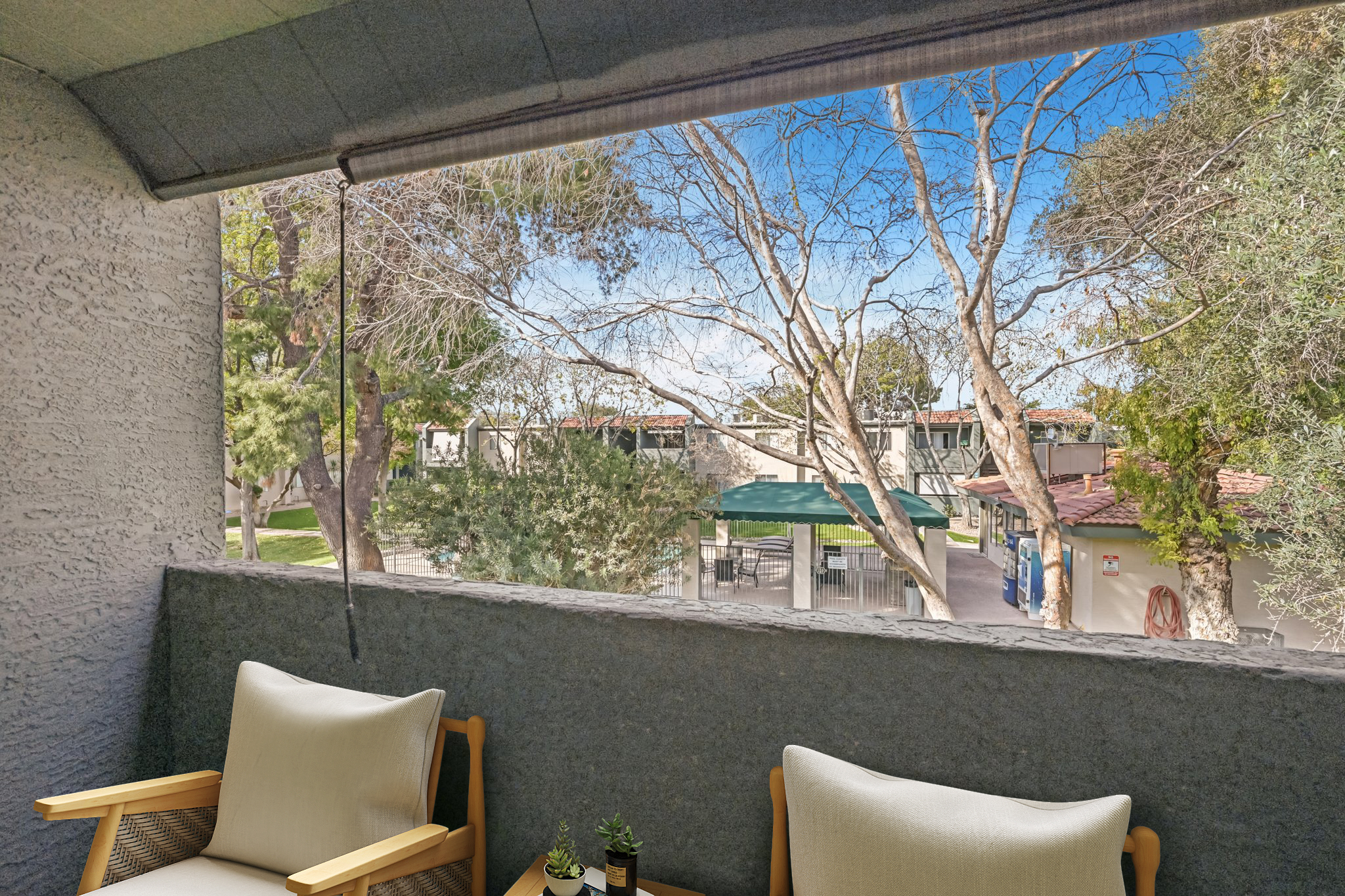 View from a balcony overlooking a green area with trees and buildings. Two light-colored chairs with small plants are placed on the balcony, creating a cozy outdoor sitting space. The sky is blue with some clouds, and there is a glimpse of a covered gathering area in the background.