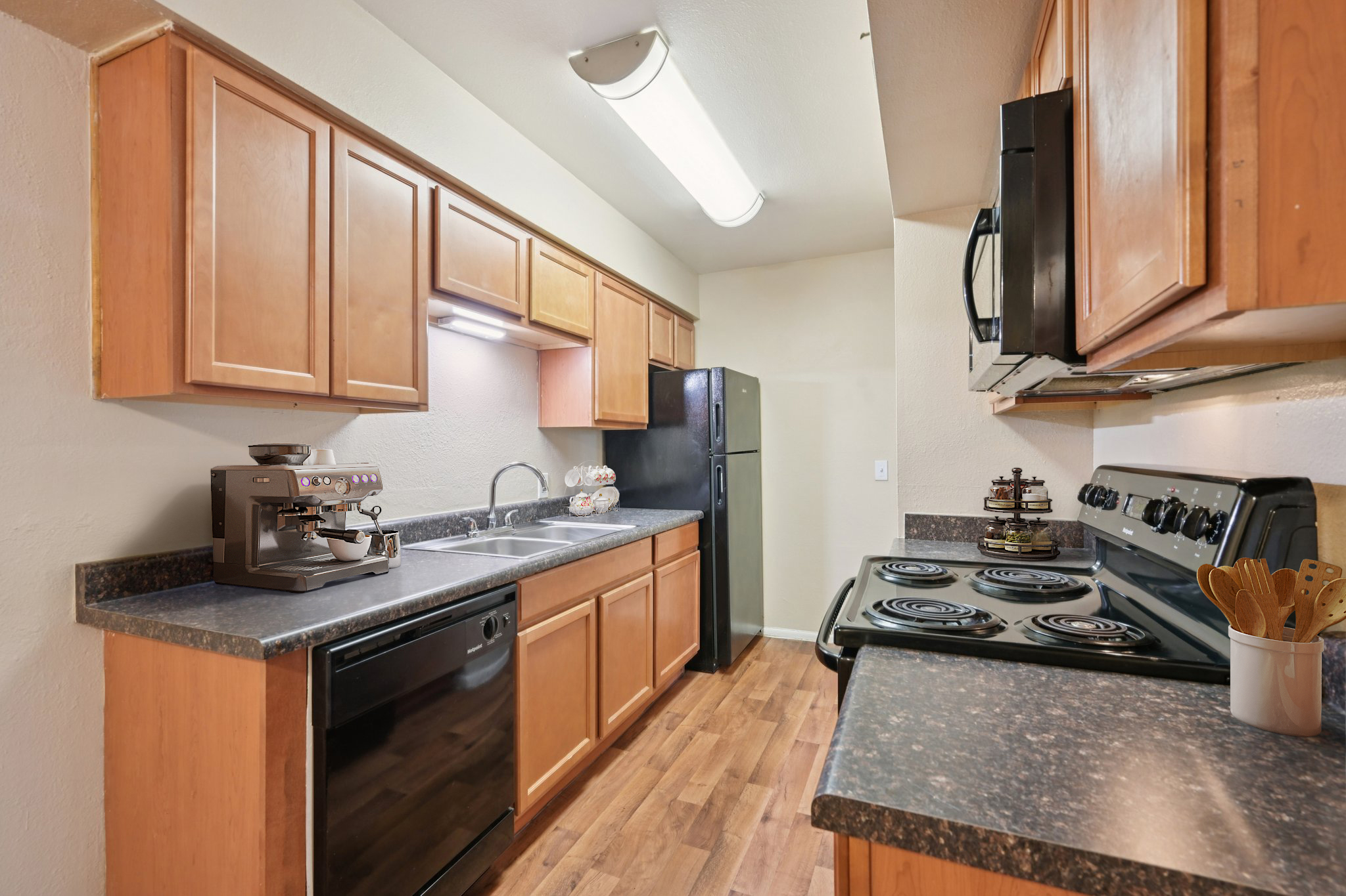 A modern kitchen featuring wooden cabinets, a countertop with kitchen utensils, a black stove, a built-in oven, and a refrigerator. Under-cabinet lighting illuminates the space, and a coffee machine is placed next to the sink. The kitchen has wooden flooring and a clean, organized appearance.