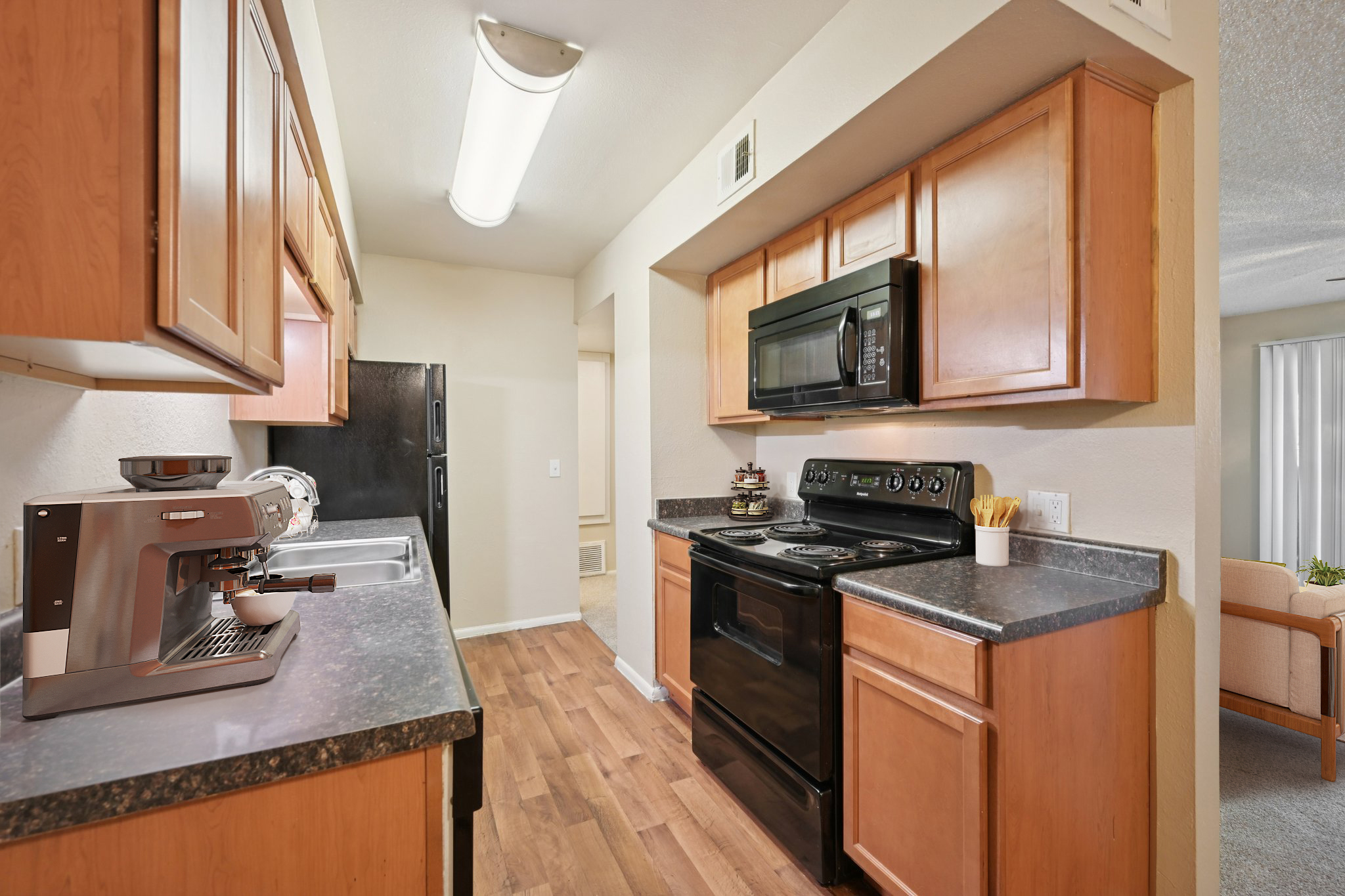 Modern kitchen featuring wooden cabinetry, black appliances including a stove and microwave, a stainless steel sink, and a coffee machine. The floor is covered with hardwood, and there's a bright light fixture overhead. Counter space is available next to the appliances.