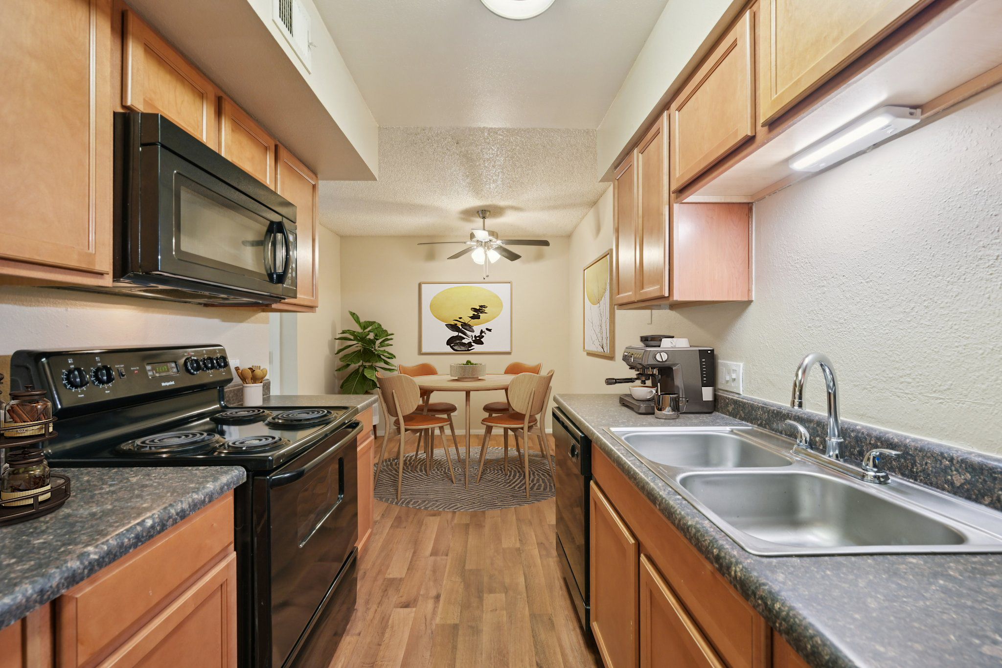 A modern kitchen featuring wooden cabinets, a black stove and microwave, and stainless steel sinks. The kitchen opens to a dining area with a round table and chairs, decorated with a large artwork and indoor plants, creating a cozy and inviting atmosphere.