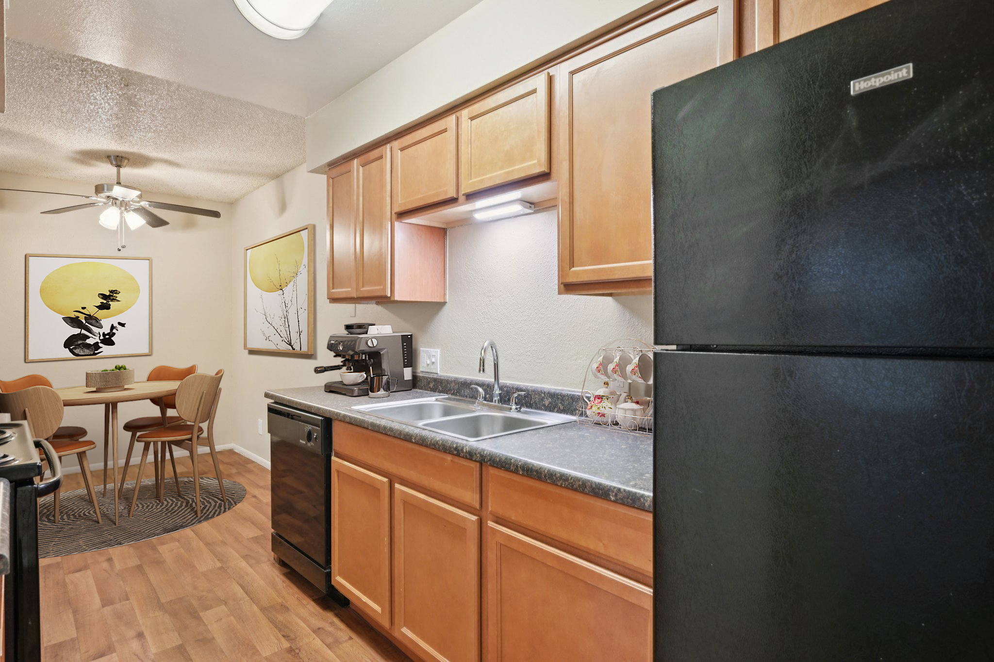 Kitchen area featuring wooden cabinetry, a black refrigerator, and a stainless steel sink. A coffee maker is positioned next to the sink. In the background, there's a dining table with four chairs and a round rug, complemented by wall artwork depicting yellow circles. A ceiling fan is visible above.