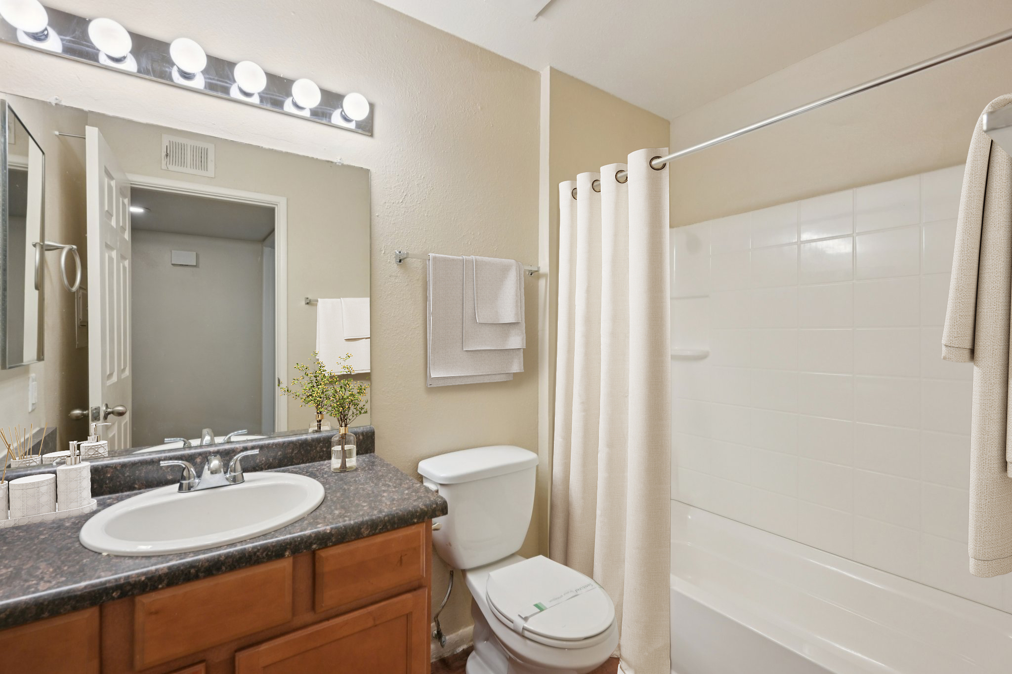A well-lit bathroom featuring a mirror above a sink with a dark countertop, a white toilet, and a shower with a white curtain. Two towels hang from a nearby rack, and a small vase with greenery sits on the counter. The walls are painted a light beige, creating a warm atmosphere.