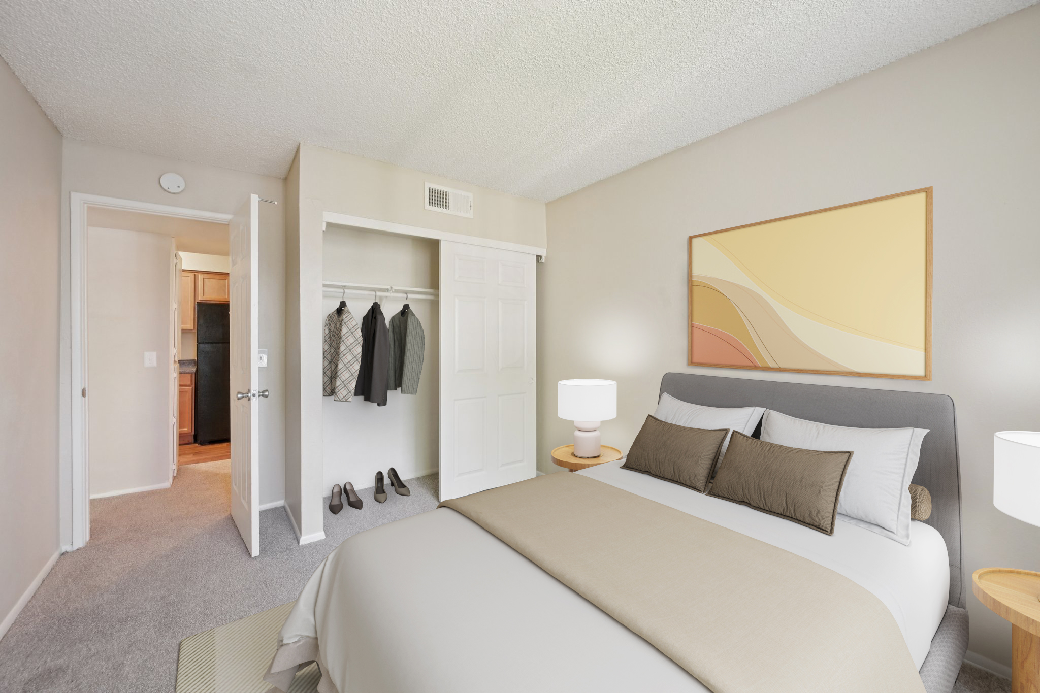 A modern bedroom featuring a neatly made bed with beige and gray bedding, two bedside tables with lamps, a closet with hanging clothes, and a doorway leading to a kitchen area. The walls are light-colored, and there is a colorful abstract painting above the bed.