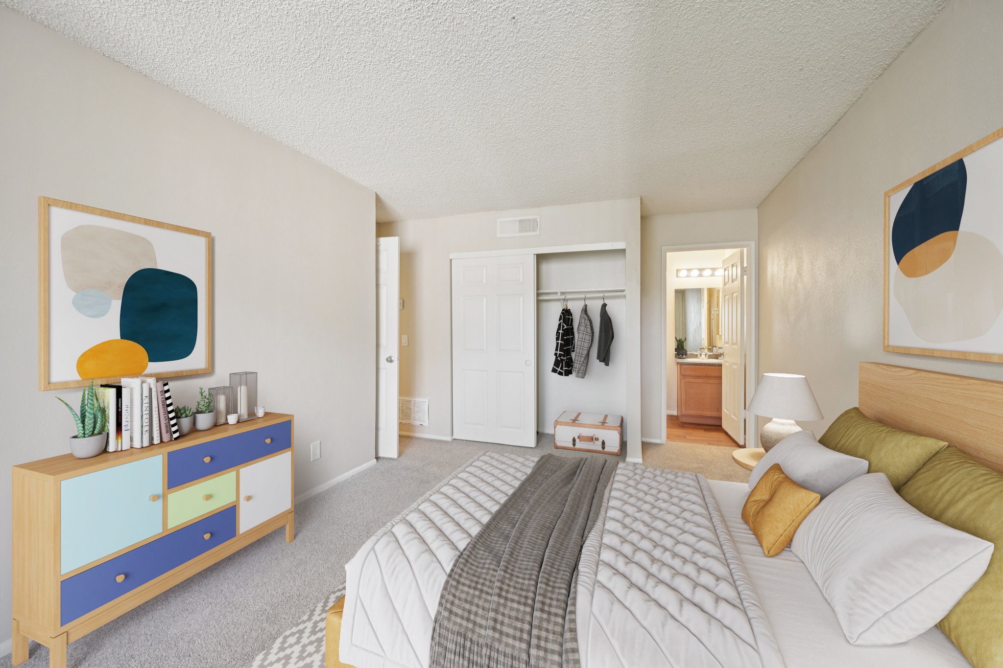 A cozy bedroom featuring a neatly made bed with decorative pillows, a colorful dresser against the wall, and modern artwork. There's a closet with hanging clothes and a bathroom visible in the background, creating a bright and inviting living space.