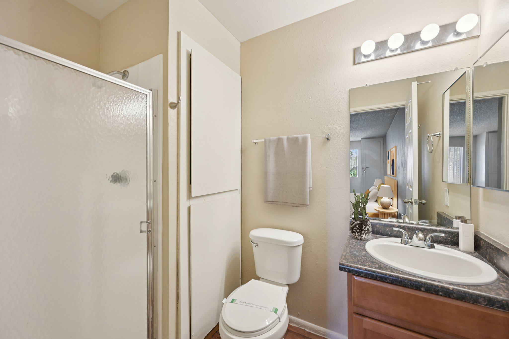 A well-lit bathroom featuring a shower stall, toilet, and sink with a mirror above. The walls are a soft beige, and there is a towel rack and small decorative elements on the counter. A light fixture with multiple bulbs adds illumination, enhancing the space's modern feel.
