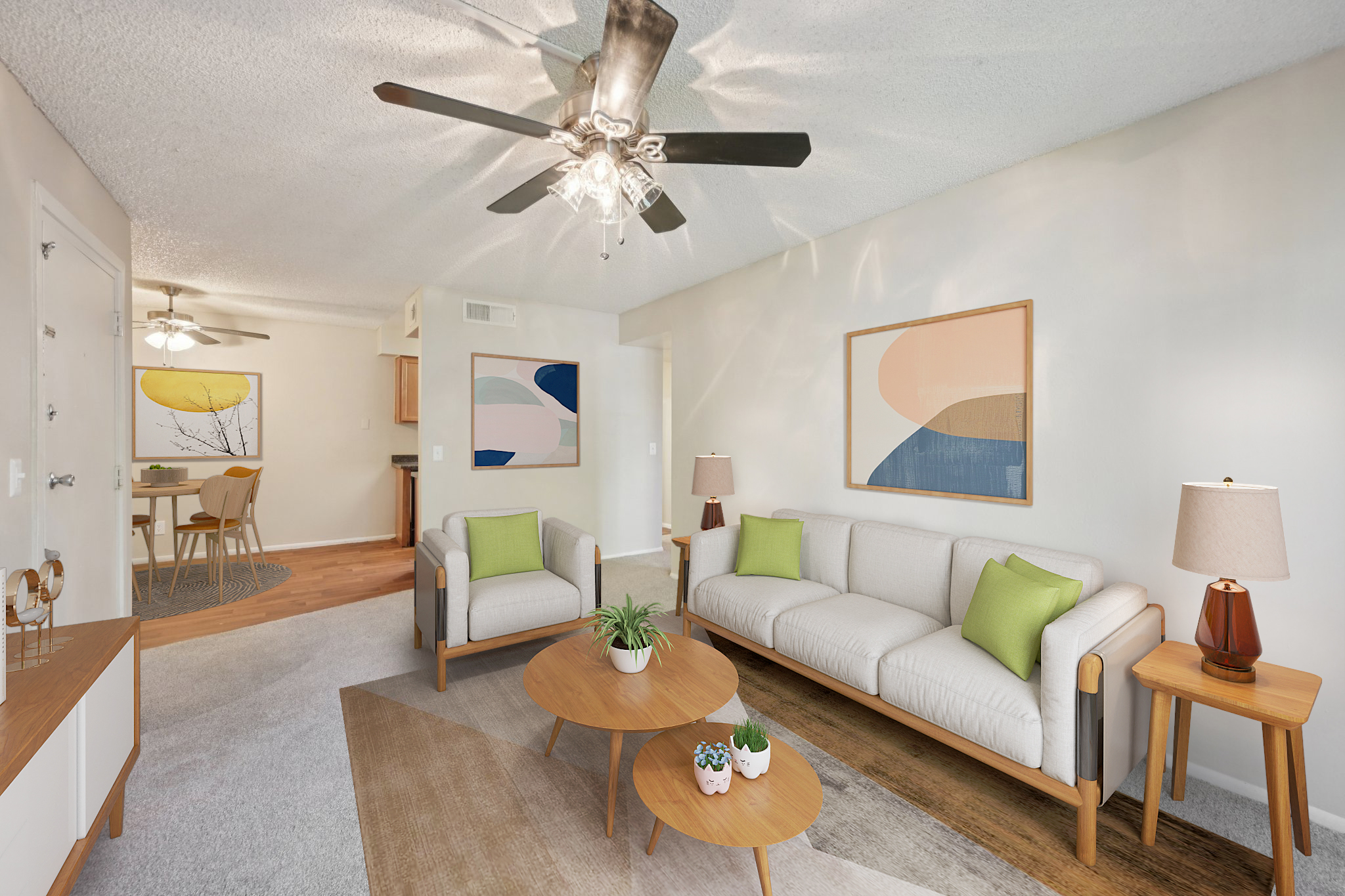 A modern living room featuring a light-colored sofa with green cushions, two round wooden coffee tables, and decorative plants. Ceiling fan with light fixtures, pastel artwork on the walls, and a large cream-colored area rug. Soft natural lighting creates a cozy atmosphere, with a dining area visible in the background.