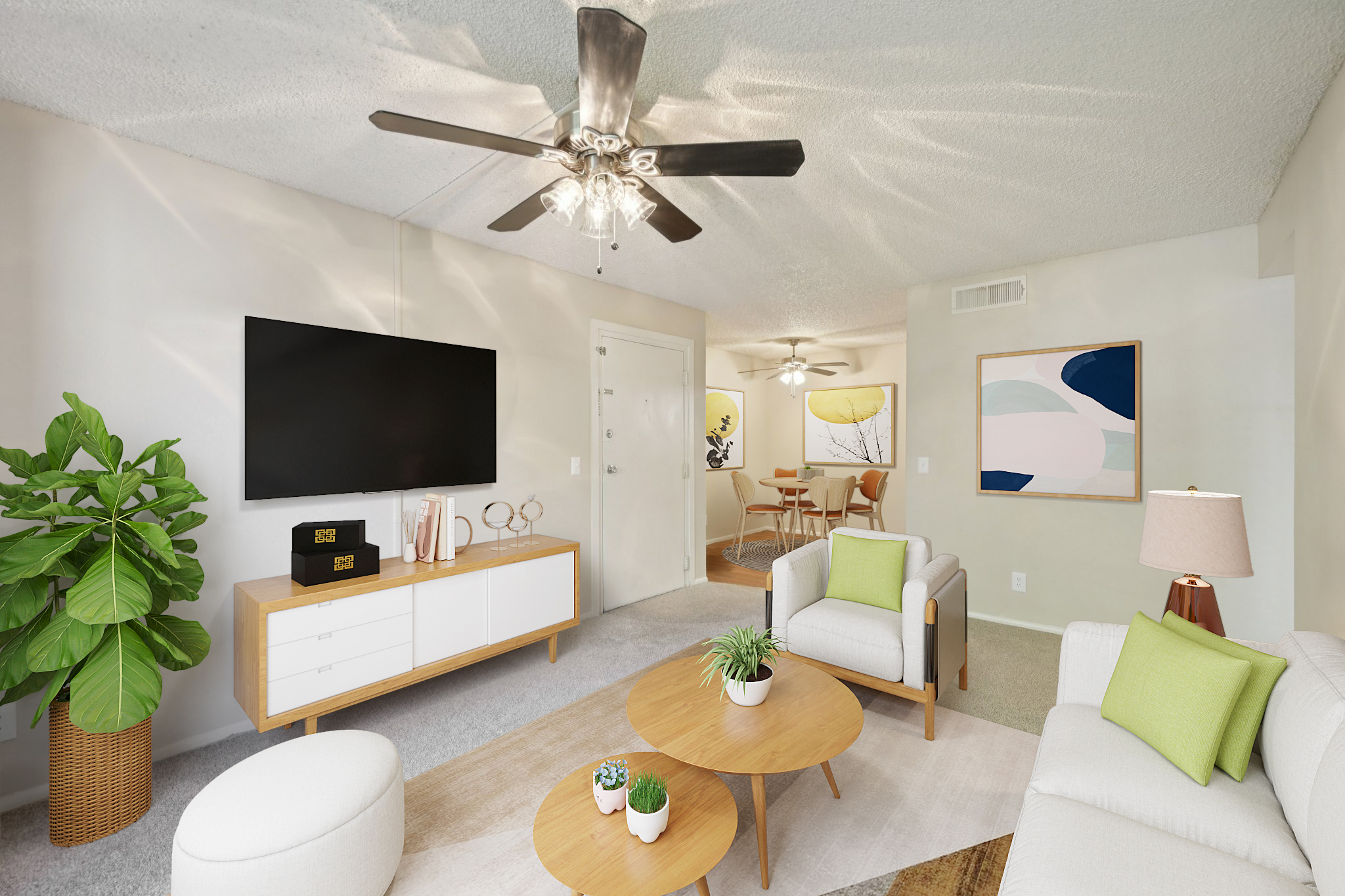 Modern living room featuring a light-colored sofa with green accent pillows, a round coffee table, and a flat-screen TV mounted on the wall. A potted plant adds a touch of greenery. In the background, there's a dining area with artwork on the wall and a round table with chairs, creating an inviting and stylish atmosphere.