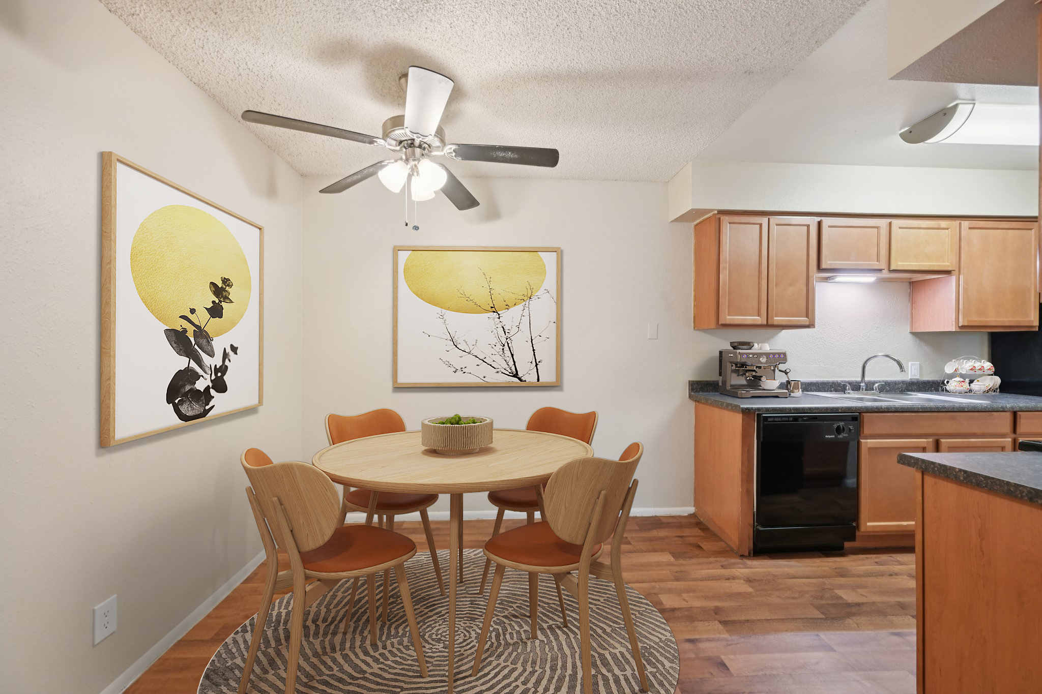 A modern kitchen and dining area featuring a round wooden table with orange chairs, two large art prints with yellow circles and nature themes on the walls, a ceiling fan, and wooden cabinetry. The space has warm lighting and a circular rug under the table.