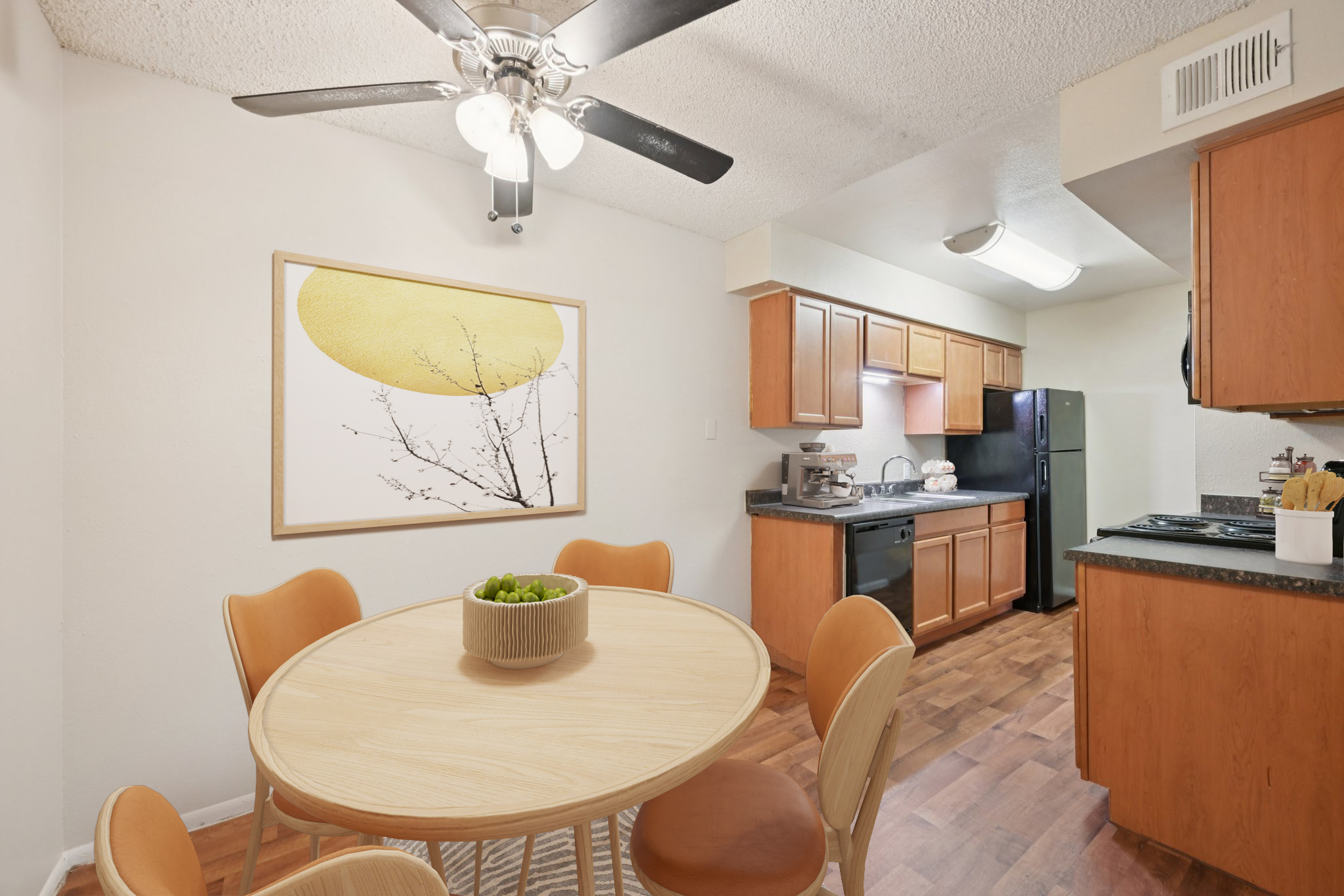 A modern kitchen and dining area featuring a round wooden table with four orange chairs, a ceiling fan, and a wall art piece depicting a minimalist design. The kitchen has wooden cabinets, a black refrigerator, and a countertop with appliances. The space is well-lit and shows a cozy, inviting atmosphere.