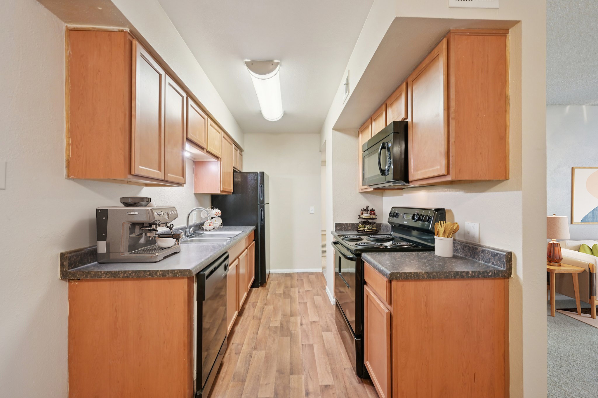 A modern kitchen with wooden cabinets and black appliances. The layout is narrow, featuring a countertop with a coffee maker and utensils. There are two black ovens, a microwave, and a refrigerator visible. The floor has wooden planks, and a light fixture is installed on the ceiling.