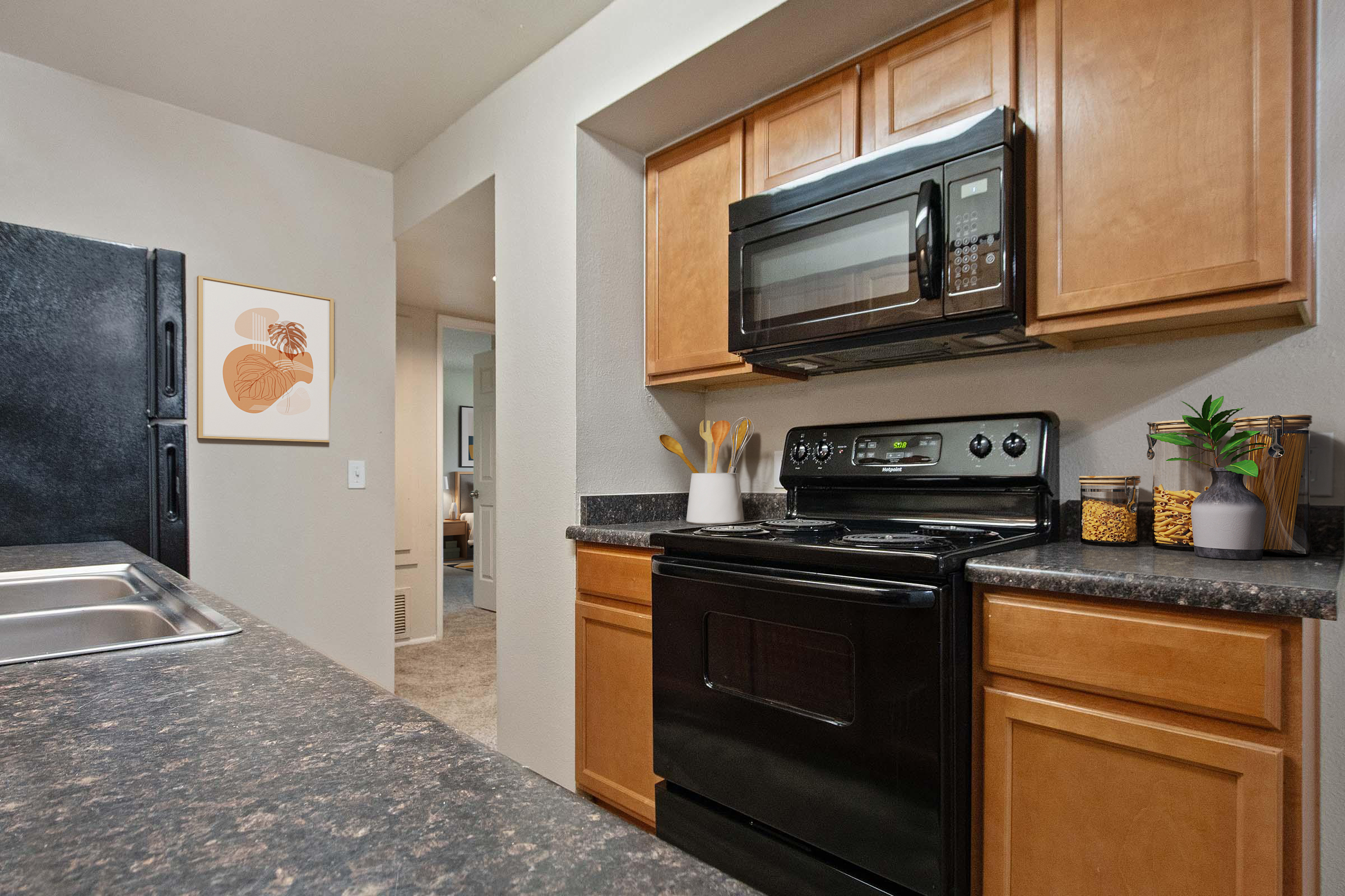 A modern kitchen featuring wooden cabinets, a black stove and microwave, and a granite countertop. There are utensils in a white container, jars of ingredients, and a framed artwork on the wall. The kitchen opens to a living area visible in the background.