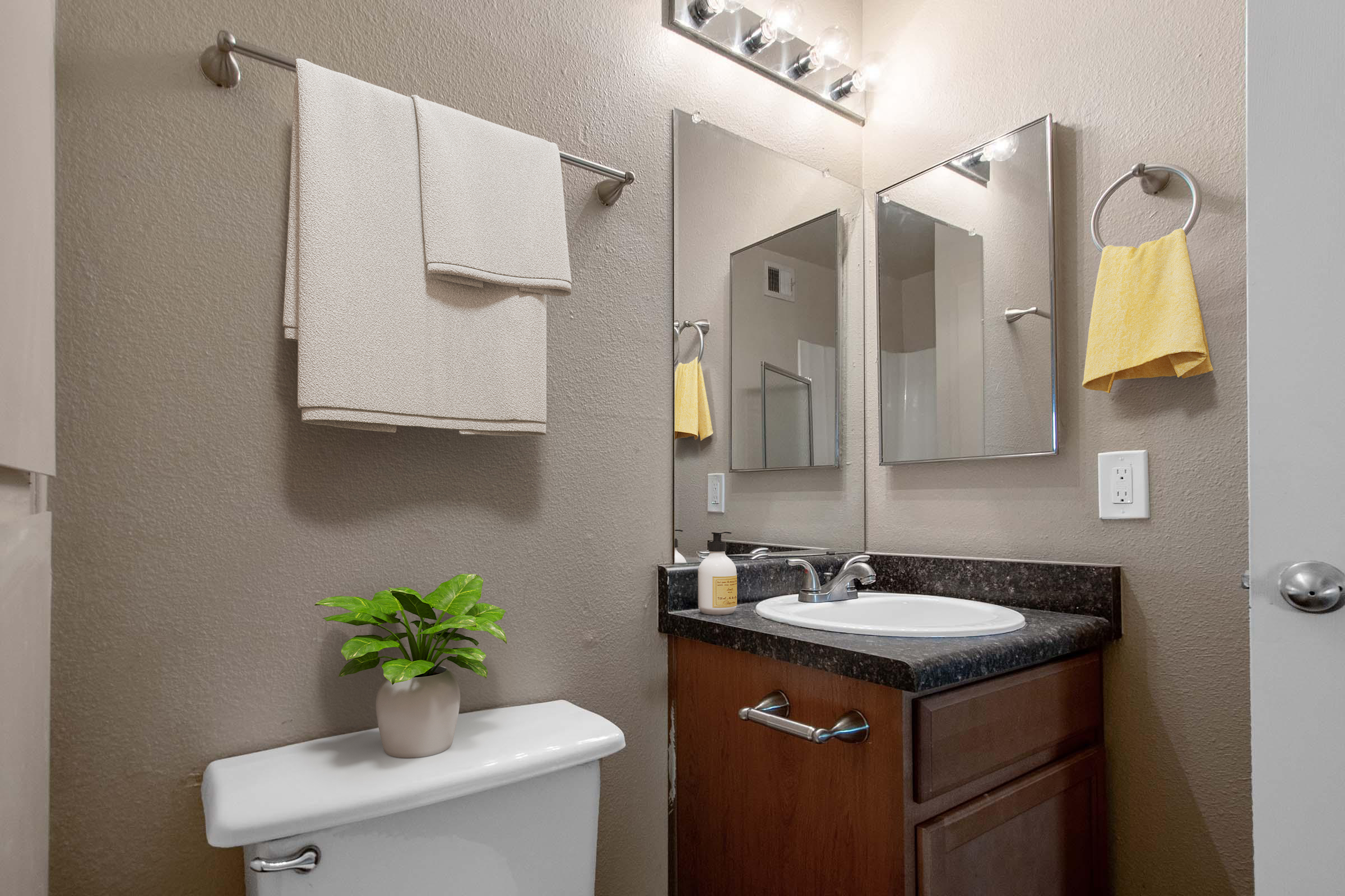 A small, modern bathroom featuring a white toilet, a sink with a dark countertop, and a wall mirror. There's a towel rack with two beige towels, a yellow hand towel, and a small green plant in a beige pot on the counter. Bright lighting illuminates the space, creating a clean and welcoming atmosphere.