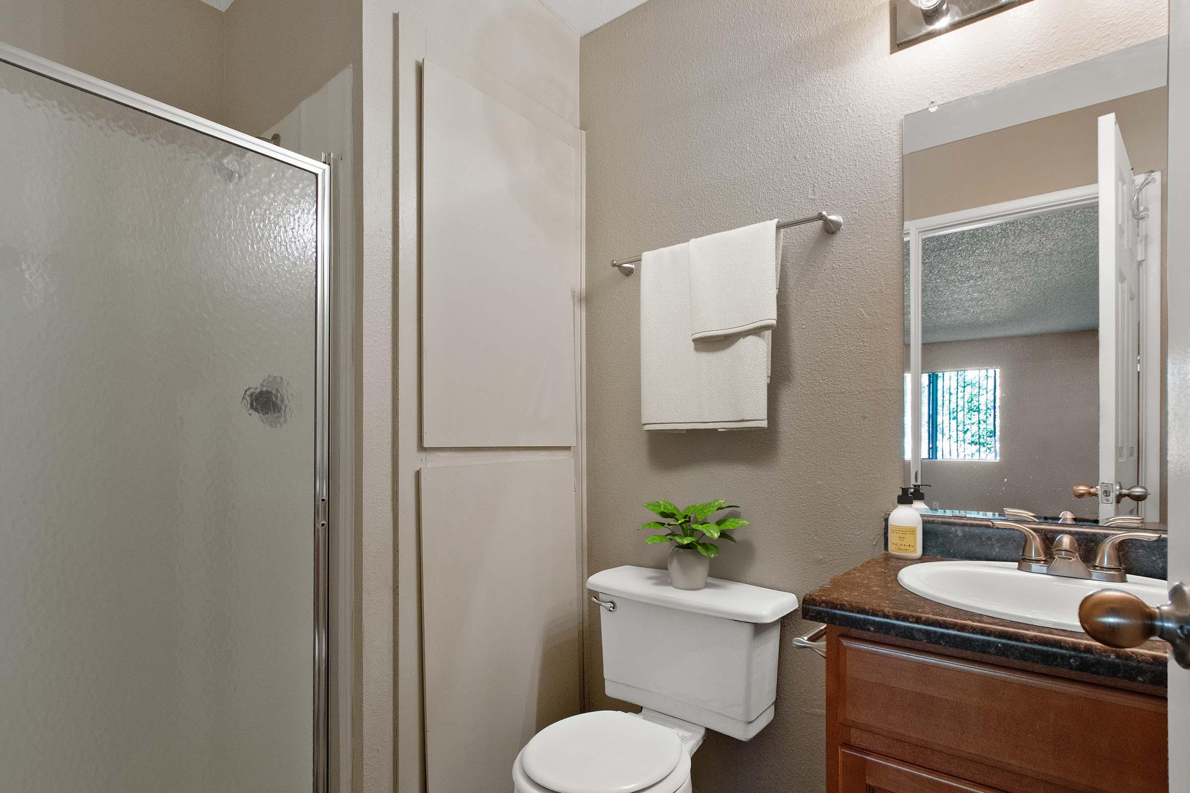 A small bathroom featuring a glass shower, a toilet, and a wooden vanity with a dark countertop. A towel hangs on a rack, and a small potted plant sits on the vanity. The walls are painted in a warm tone, and there is a mirror above the sink reflecting light from a window.