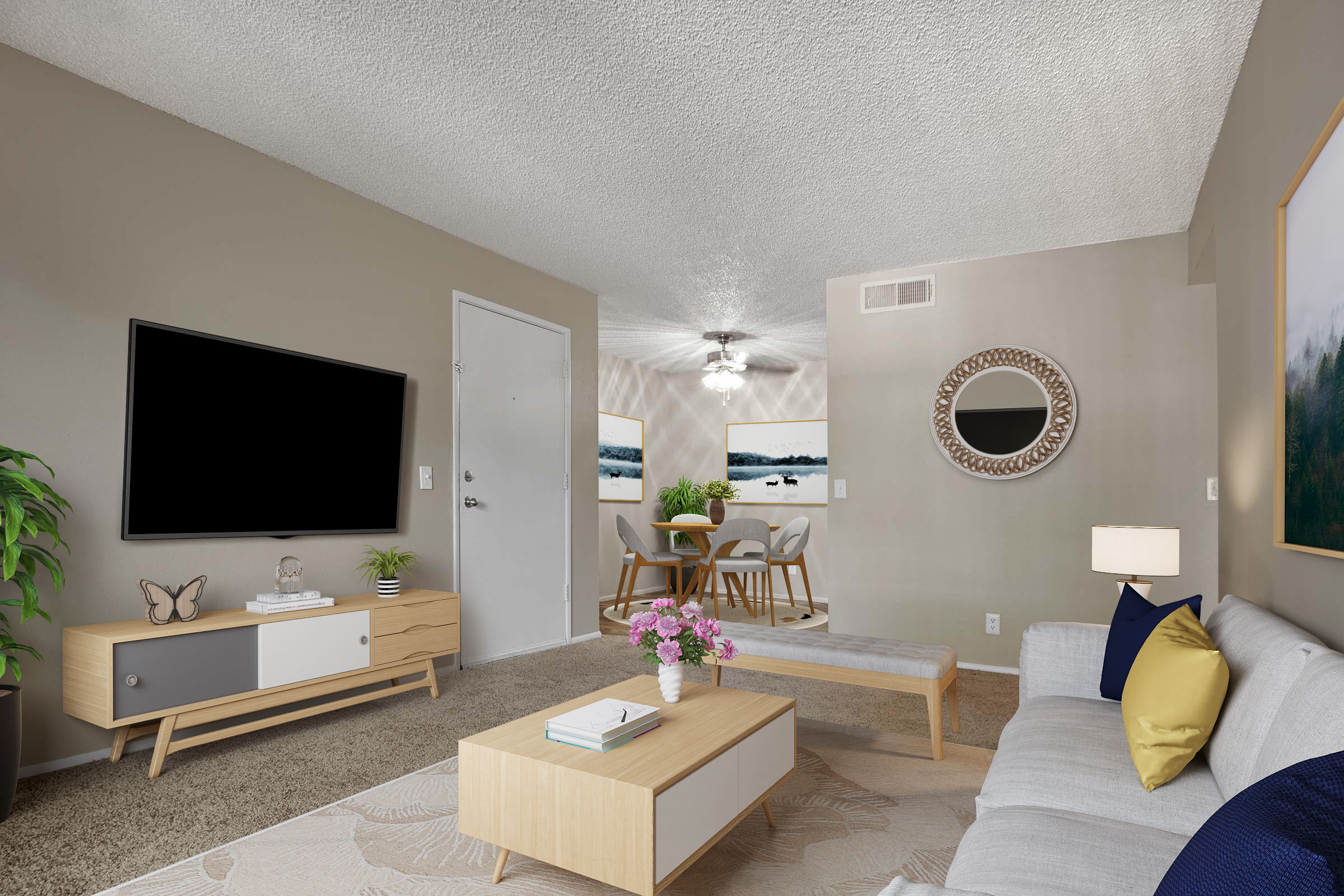 Modern living room with a light-colored sofa, a wooden coffee table, and a TV mounted on the wall. A plant adds greenery, and there is a small vase with flowers on the table. In the background, a dining area features a circular table surrounded by chairs, with a ceiling fan and a mirror on the wall.