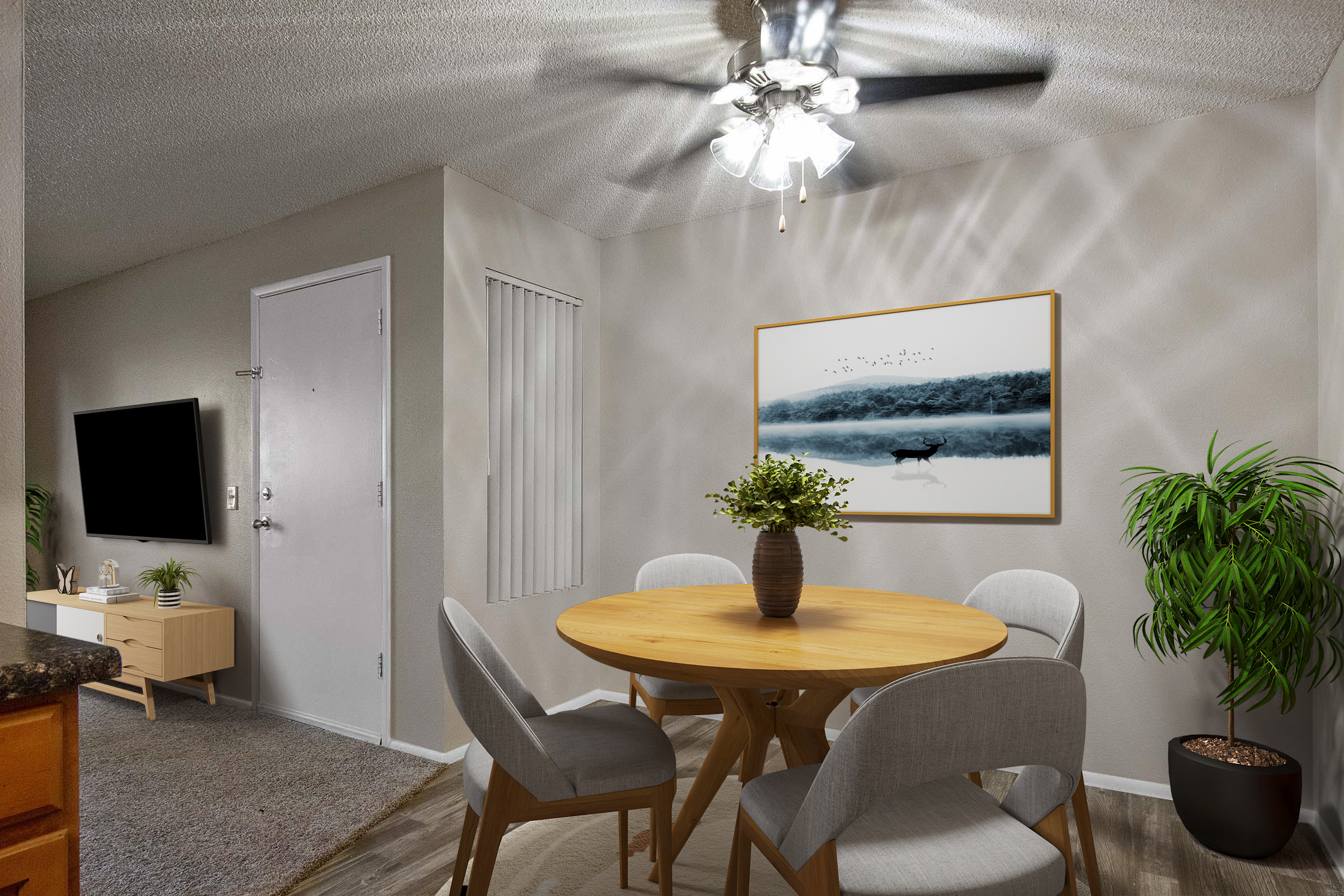 A cozy dining area featuring a round wooden table surrounded by four light-colored chairs. A small potted plant adorns the table, and a large framed picture of a serene lake scene hangs on the wall. A ceiling fan with lights and a flat-screen TV are also visible in the room, creating an inviting atmosphere.