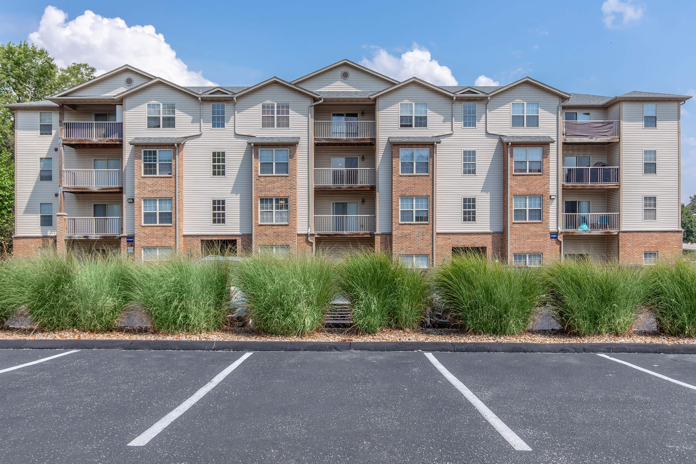 A multi-story residential building featuring a mix of brick and siding exteriors, with balconies on each floor. In the foreground, there is a neatly landscaped area with tall grass plants, and a parking lot with marked spaces beneath a clear blue sky.