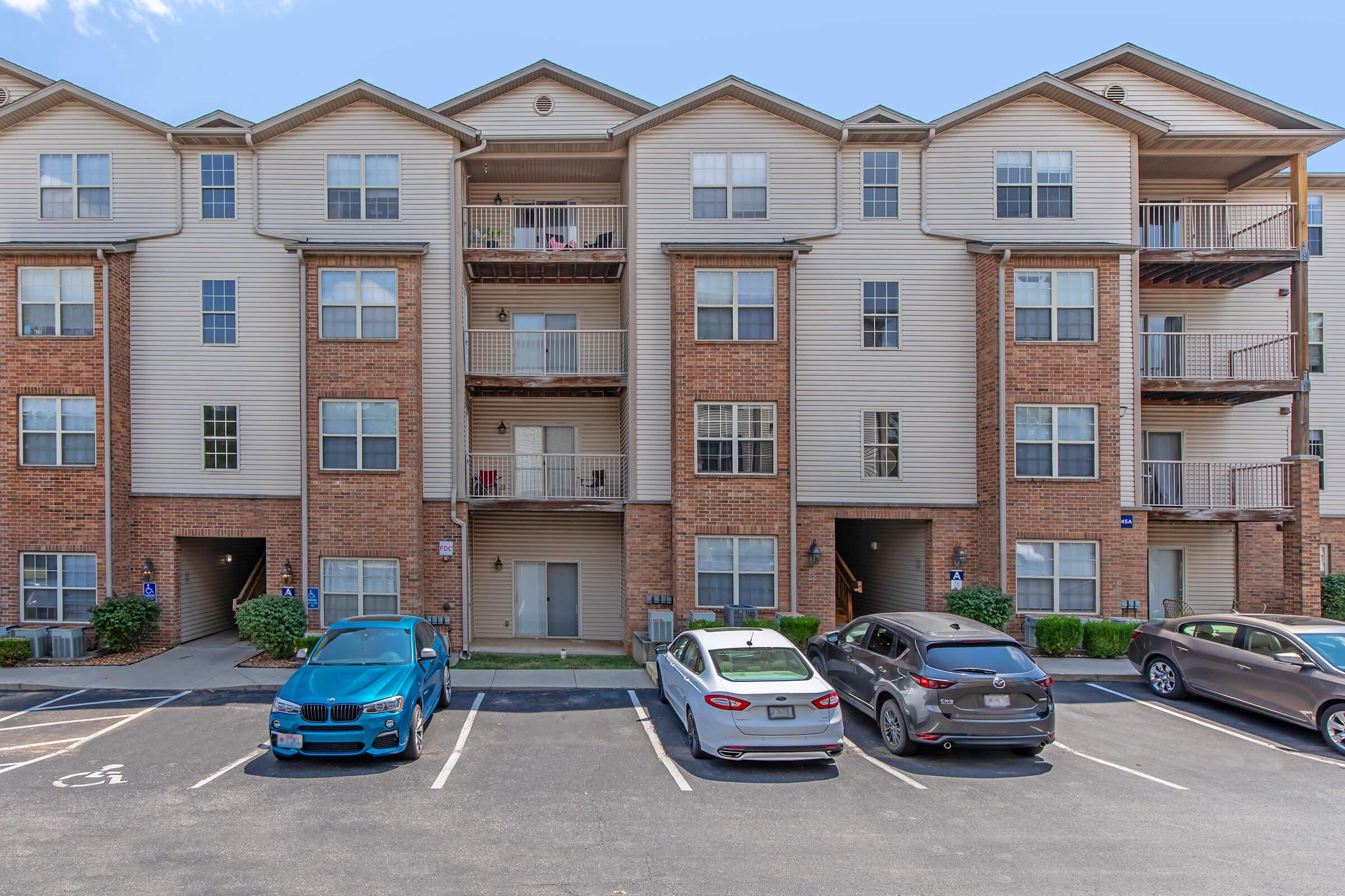 A multi-story residential apartment building featuring brick and siding exteriors. Several parking spaces are visible in front, occupied by different colored cars. The building has balconies and landscaped areas with greenery. Clear blue skies create a bright setting.