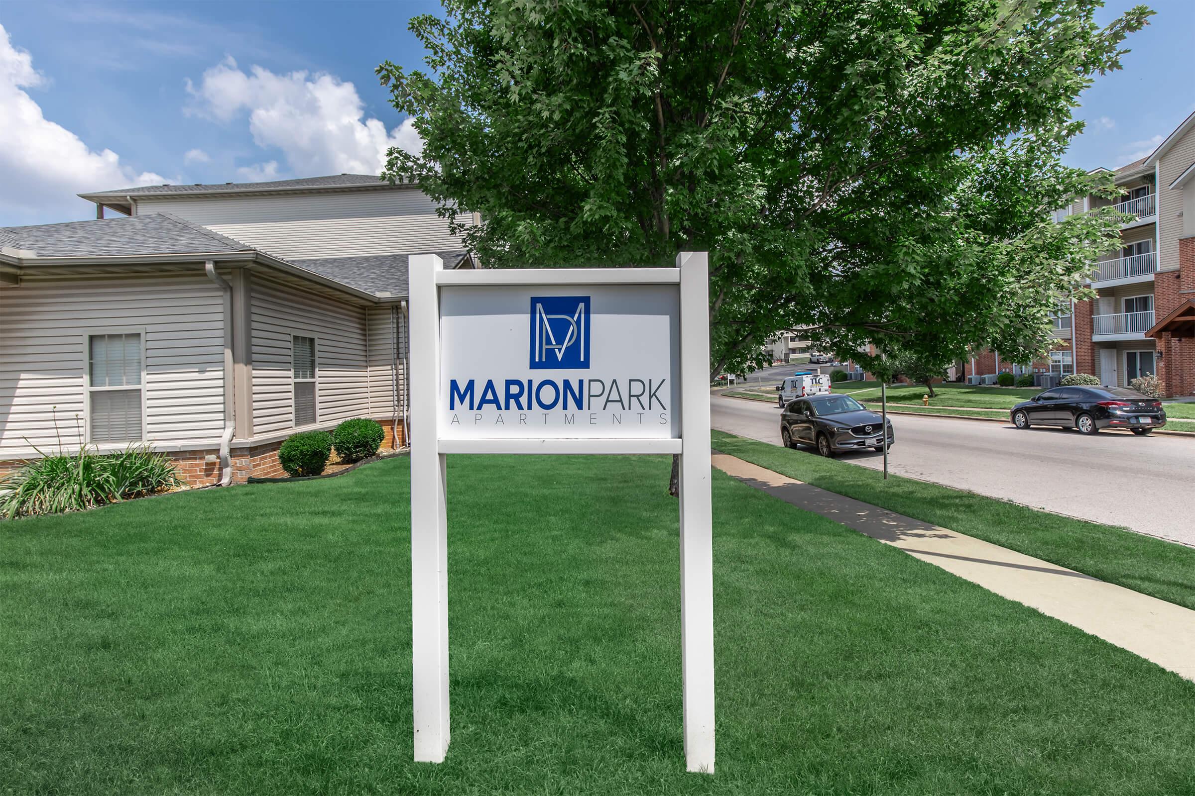 Sign for Marion Park Apartments, displayed on a white post in a well-maintained grassy area. In the background, there are residential buildings and trees, with a clear blue sky above. A car is parked along the street.