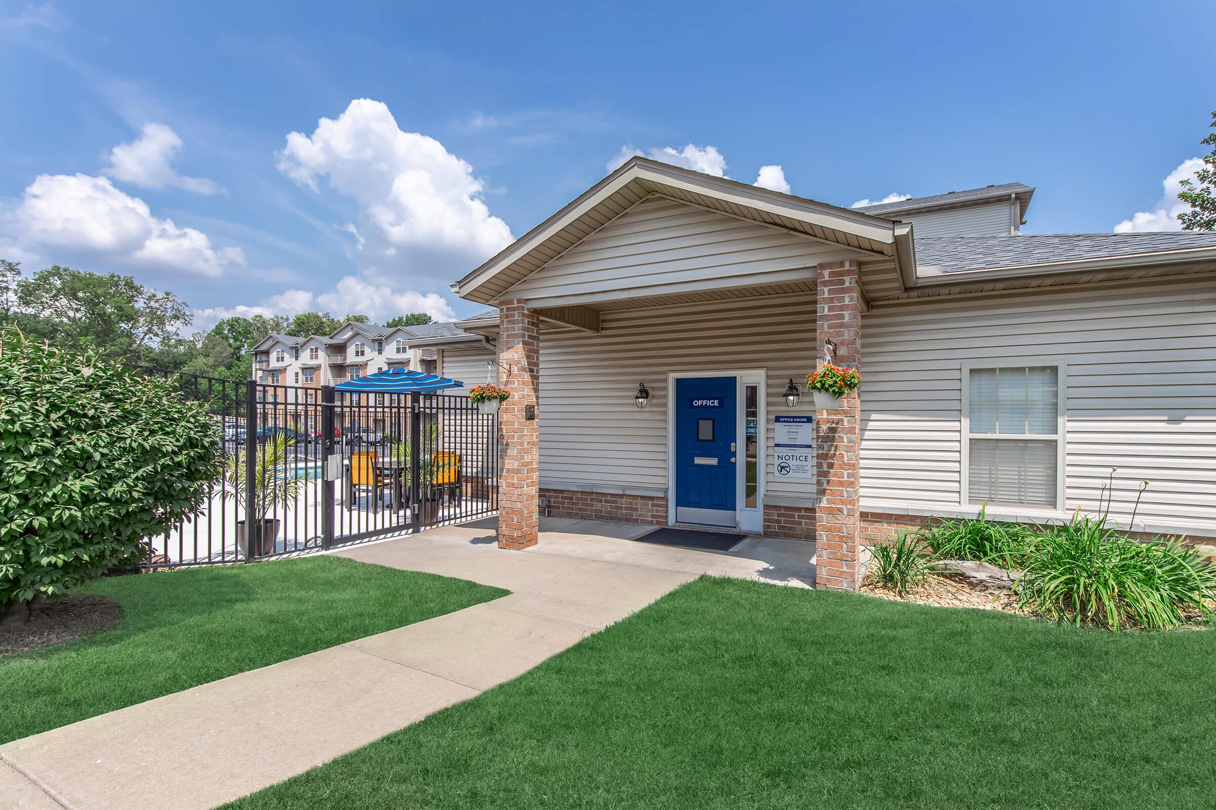 Exterior view of a residential office building with a blue door and stone accents, surrounded by green grass and landscaping. In the background, a fenced swimming pool area is visible, with apartment buildings beyond. Bright, sunny skies and fluffy clouds create a cheerful atmosphere.