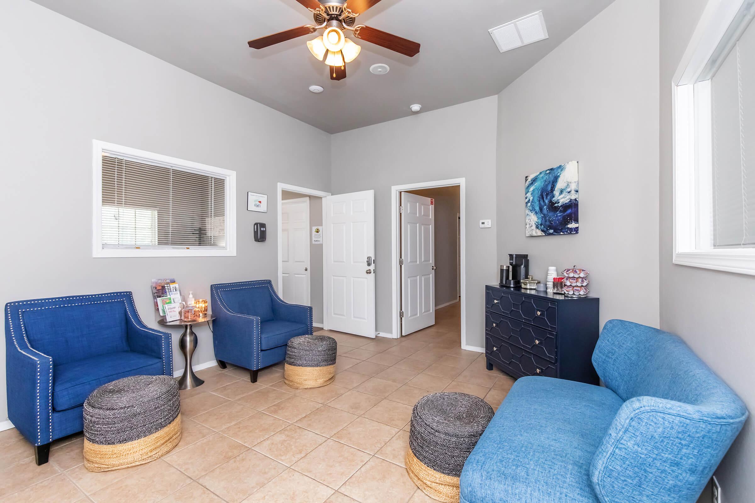 A welcoming waiting area featuring two blue armchairs, a blue sofa, and two round ottomans. The walls are light gray, and there is a small side table with a candle. A decorative piece hangs on one wall, while a dark cabinet is visible on another. Natural light filters in through a window.