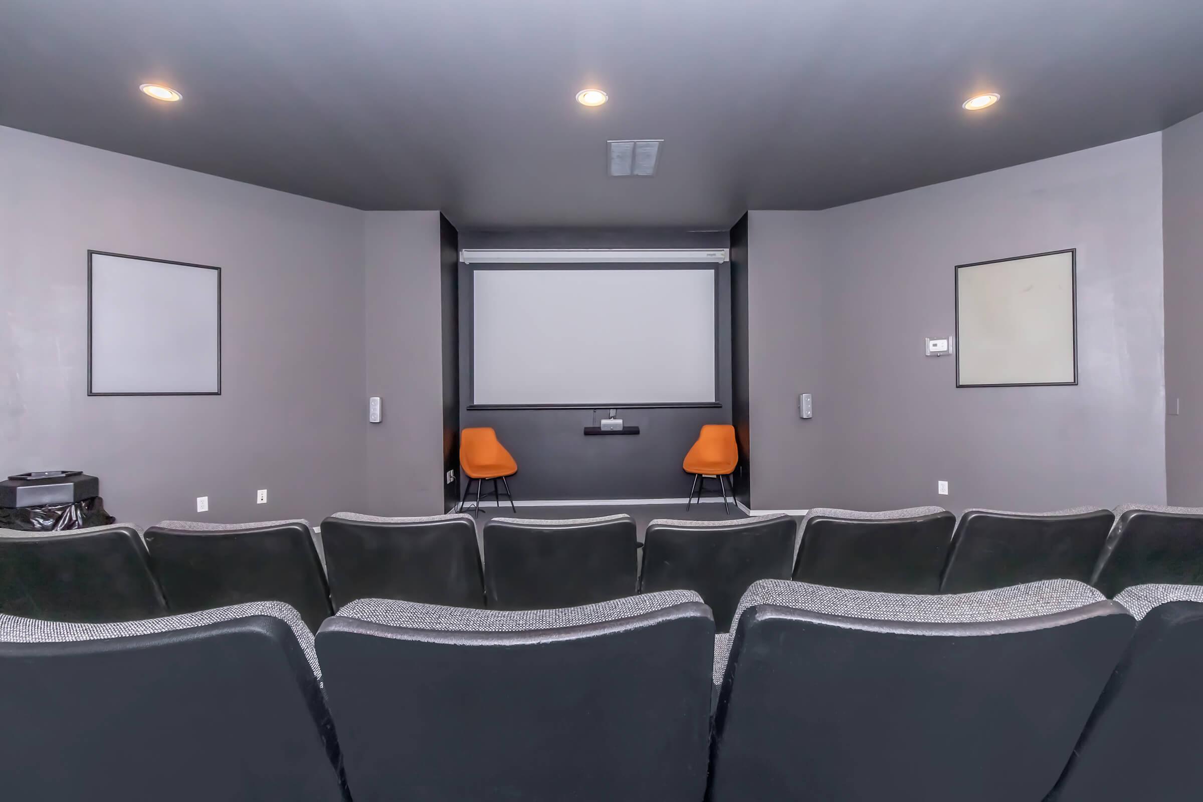 A view of a small home theater or screening room featuring a large projection screen at the front, two orange chairs positioned in front of it, and tiered black seating arranged in rows. The walls are painted a neutral gray, and there are empty frames on either side of the screen.