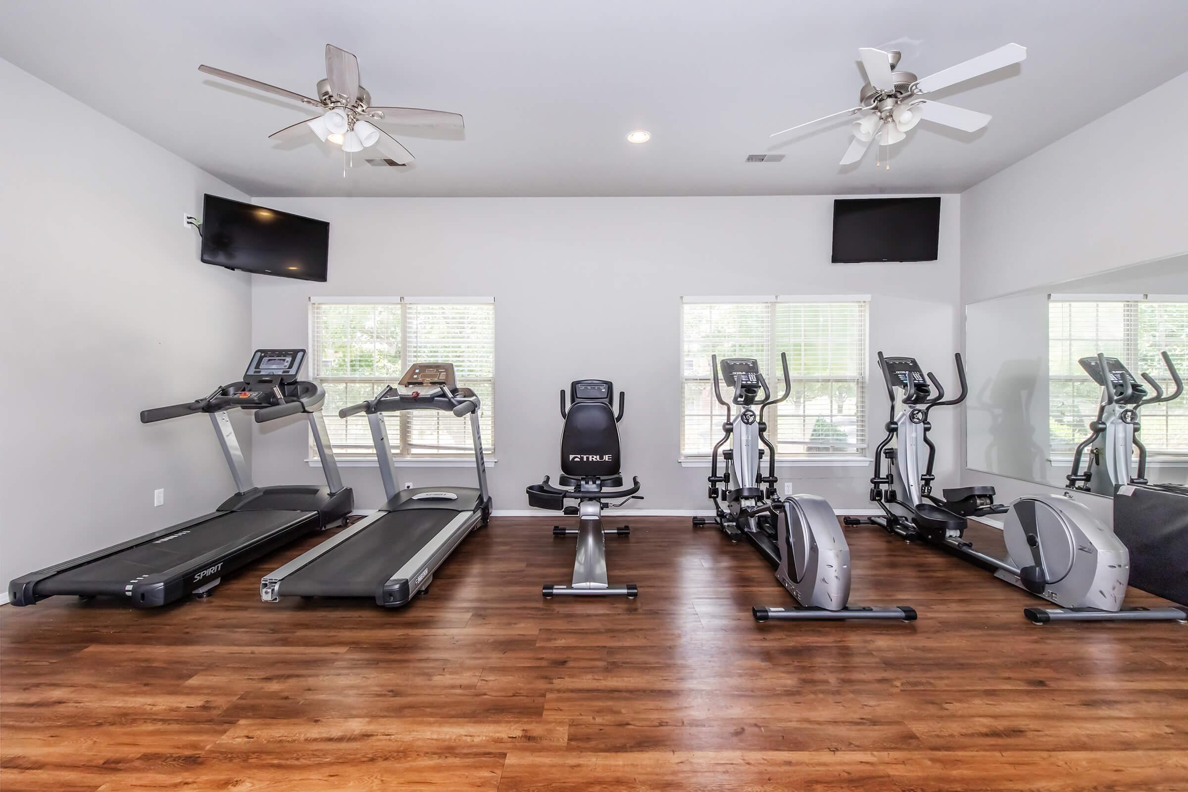 A modern home gym featuring multiple exercise machines, including a treadmill, an elliptical, and a stationary bike. The room has wooden flooring, ceiling fans, large windows letting in natural light, and mirrors on one wall. Two flat-screen TVs are mounted on the walls for entertainment.