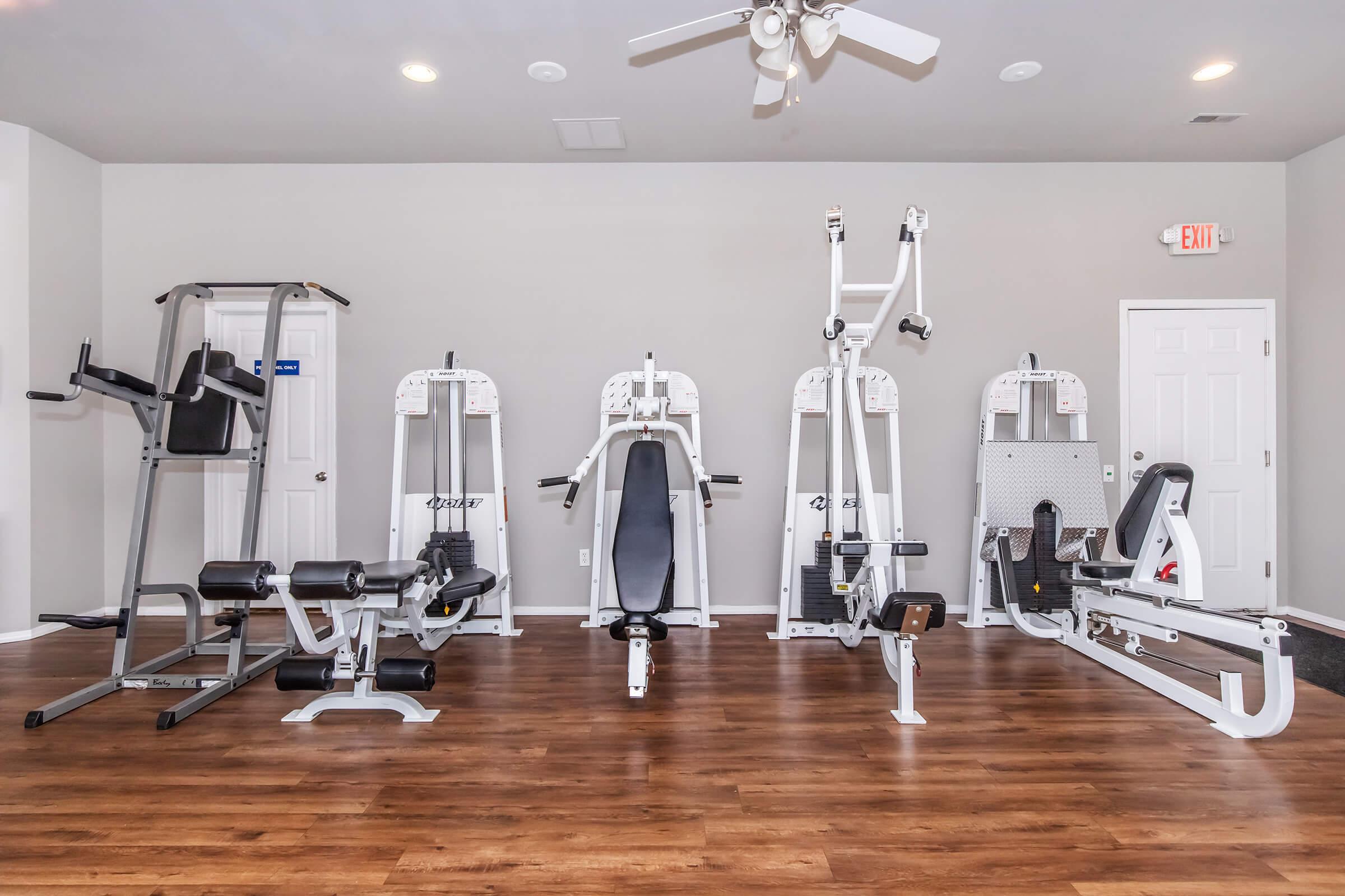 A well-equipped home gym featuring various exercise machines including a weight bench, multi-station gym, rowing machine, and a pull-up bar. The space has a wooden floor, ceiling fan, and neutral-colored walls, creating a bright and inviting workout environment.