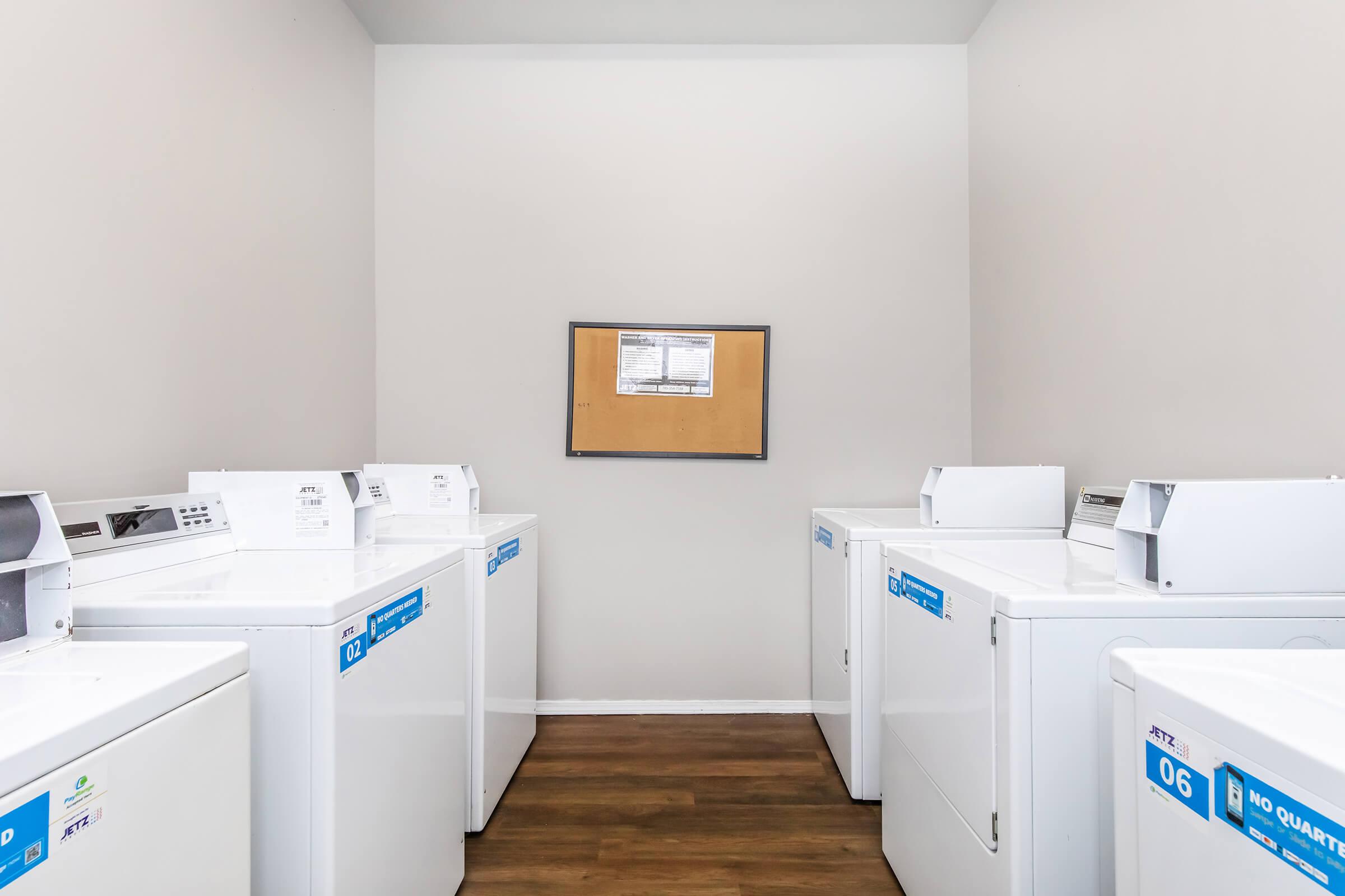 A laundry room featuring several white washing machines and dryers lined up against the walls. There is a bulletin board on the wall with a notice, and the floor is wood-like. The room has neutral-colored walls and ample space.