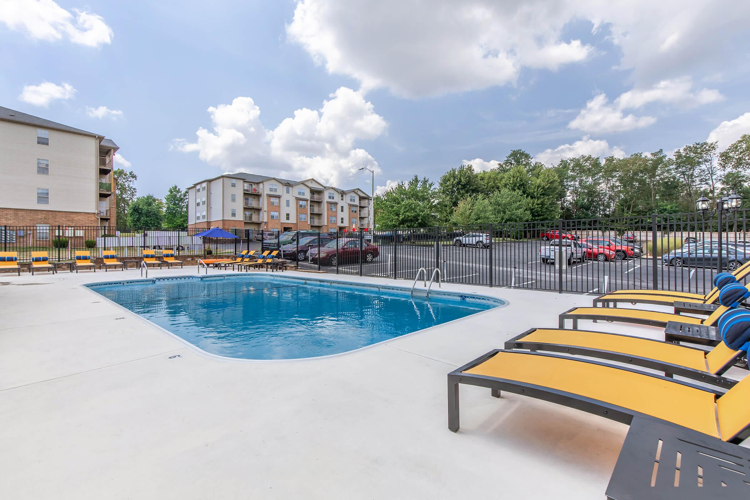 A spacious outdoor swimming pool surrounded by lounge chairs with yellow cushions. The pool area is enclosed by a black fence, and in the background, there are several apartment buildings and parked cars. The sky is partly cloudy, adding to the inviting atmosphere of the scene.