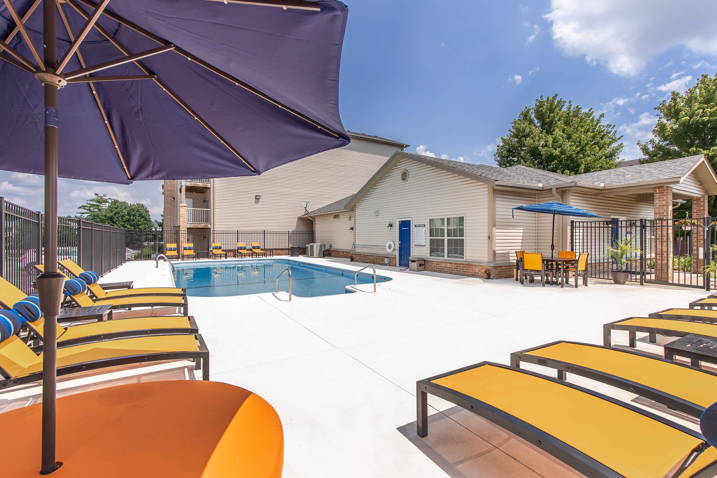 A bright outdoor swimming pool area featuring lounge chairs with yellow cushions and a large purple umbrella. The pool is surrounded by a fenced area with trees in the background. A blue table and chairs are also visible, with a well-maintained building nearby under a clear blue sky.