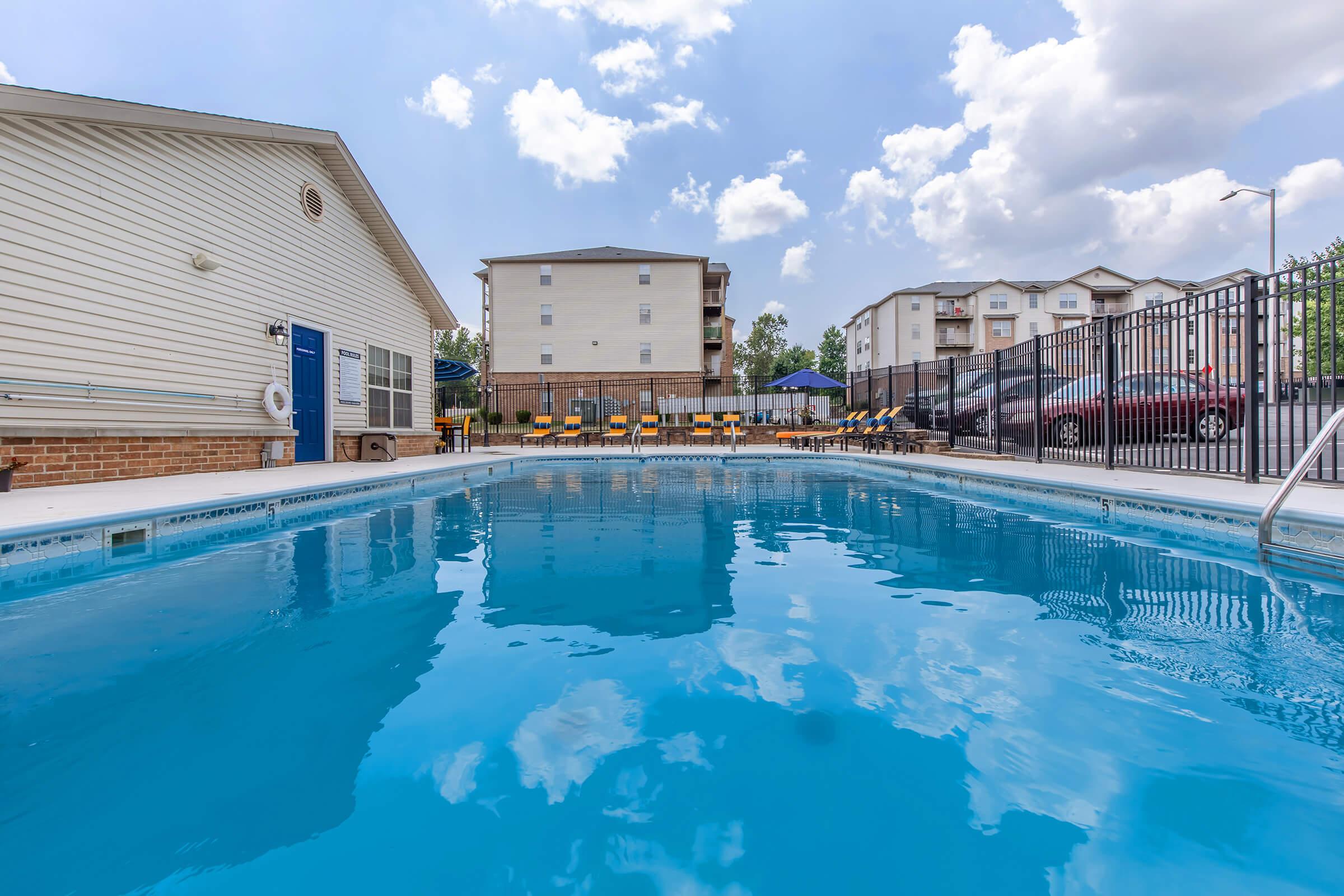 A clear blue swimming pool with steps leading in, surrounded by a black fence. In the background, there are apartment buildings and a sunny sky with scattered clouds. Lounge chairs are positioned near the poolside, creating a relaxing atmosphere.