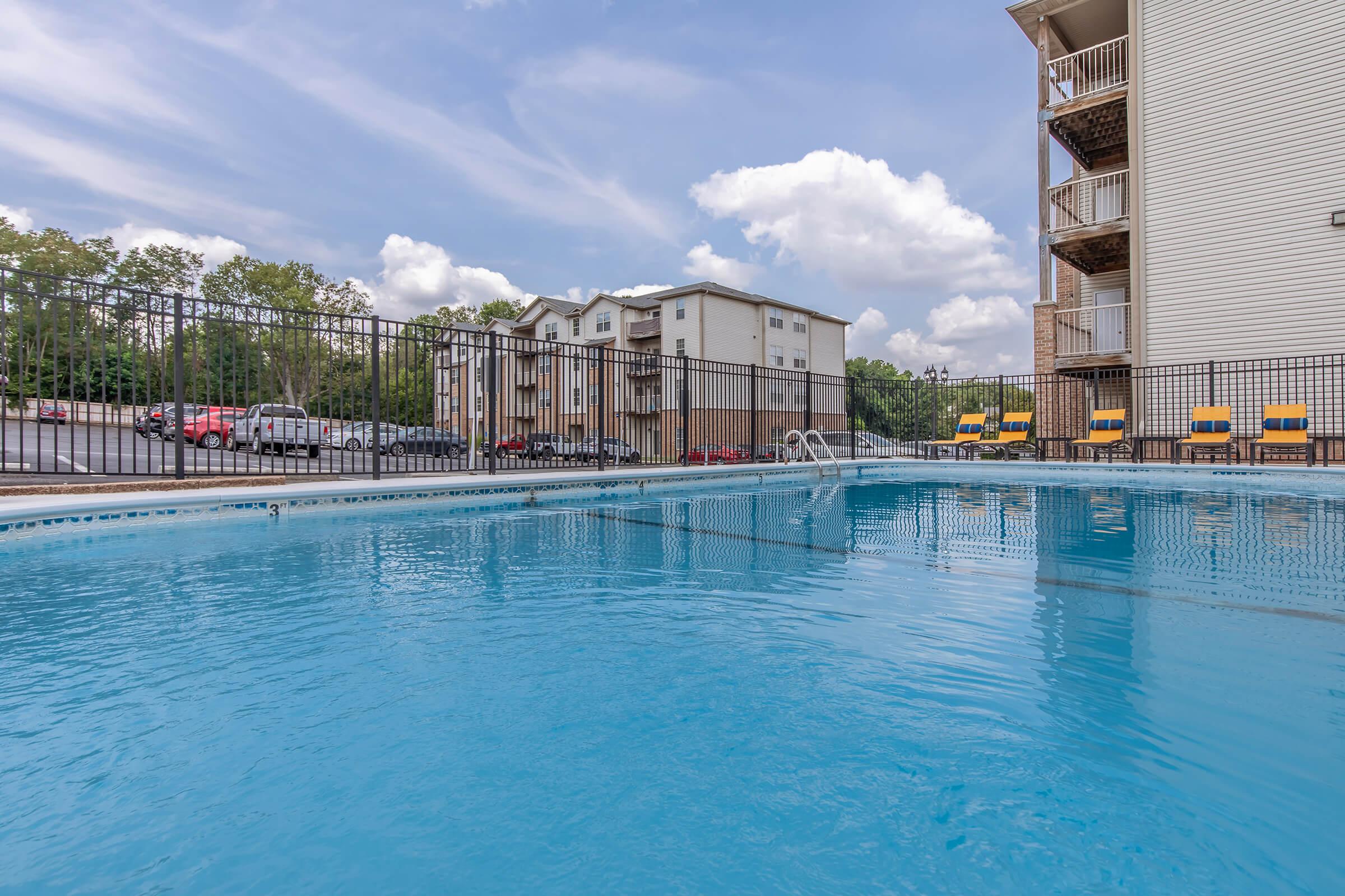 A clear swimming pool surrounded by a black metal fence, with bright blue water reflecting the sky. In the background, there are multi-story apartment buildings and a parking area with several cars. The scene is set under a partly cloudy sky, creating a serene and inviting atmosphere.