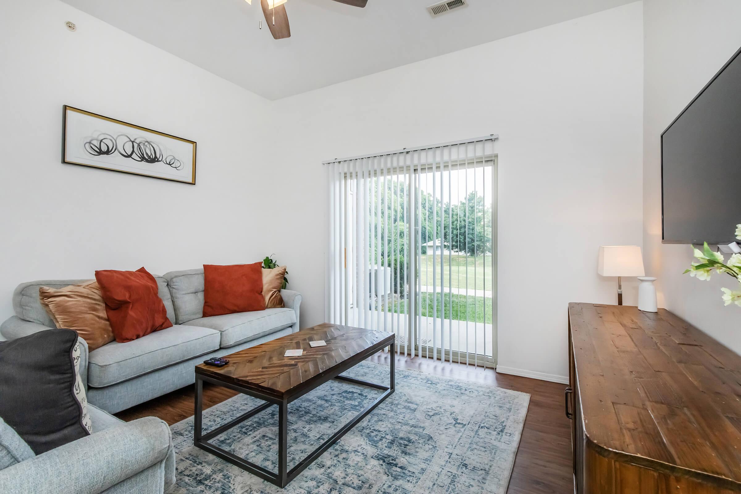 A bright living room featuring a light blue sofa with orange cushions, a wooden coffee table, and a flat-screen TV mounted on the wall. Large glass sliding doors offer a view of greenery outside, while a patterned area rug adds warmth to the space. A lamp and decorative artwork complete the cozy atmosphere.