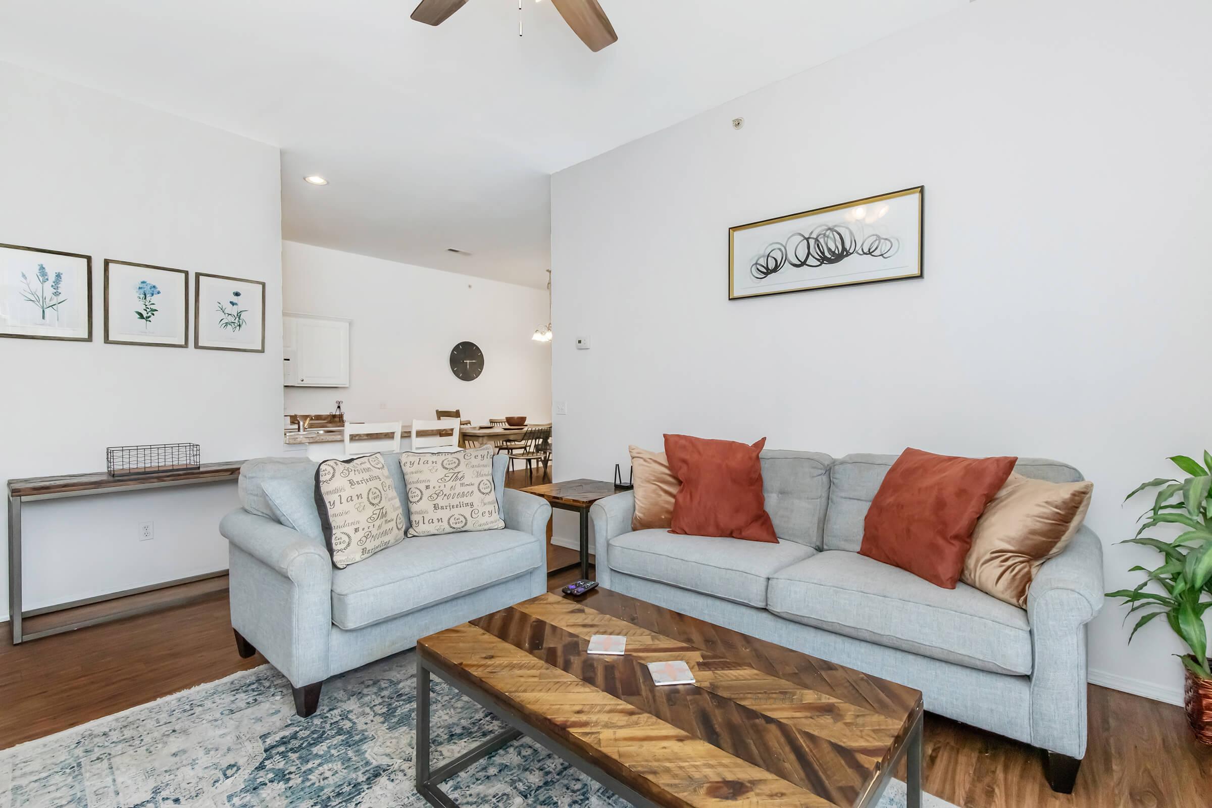 A cozy living room featuring a light gray sofa with decorative pillows, a wooden coffee table, and framed wall art. The space has wooden flooring and a ceiling fan, with a dining area visible in the background, showcasing a round mirror and additional seating.