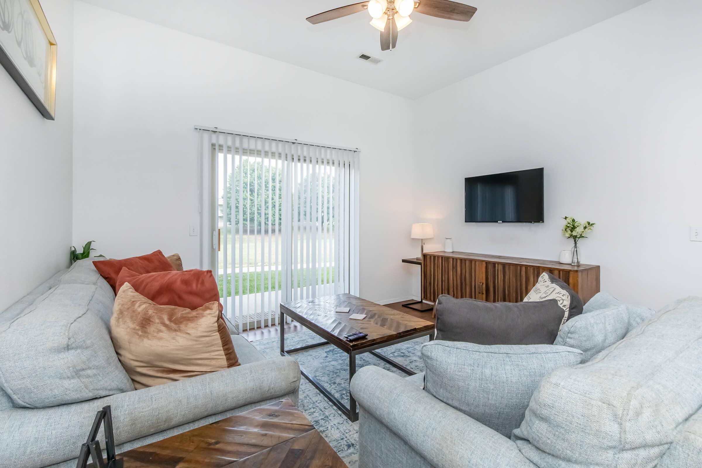 A cozy living room featuring two comfortable sofas with decorative pillows, a wooden coffee table, and a media console with a TV. A sliding glass door with vertical blinds brings in natural light, overlooking a lush green area outside. The walls are painted white, enhancing the airy feel of the space.