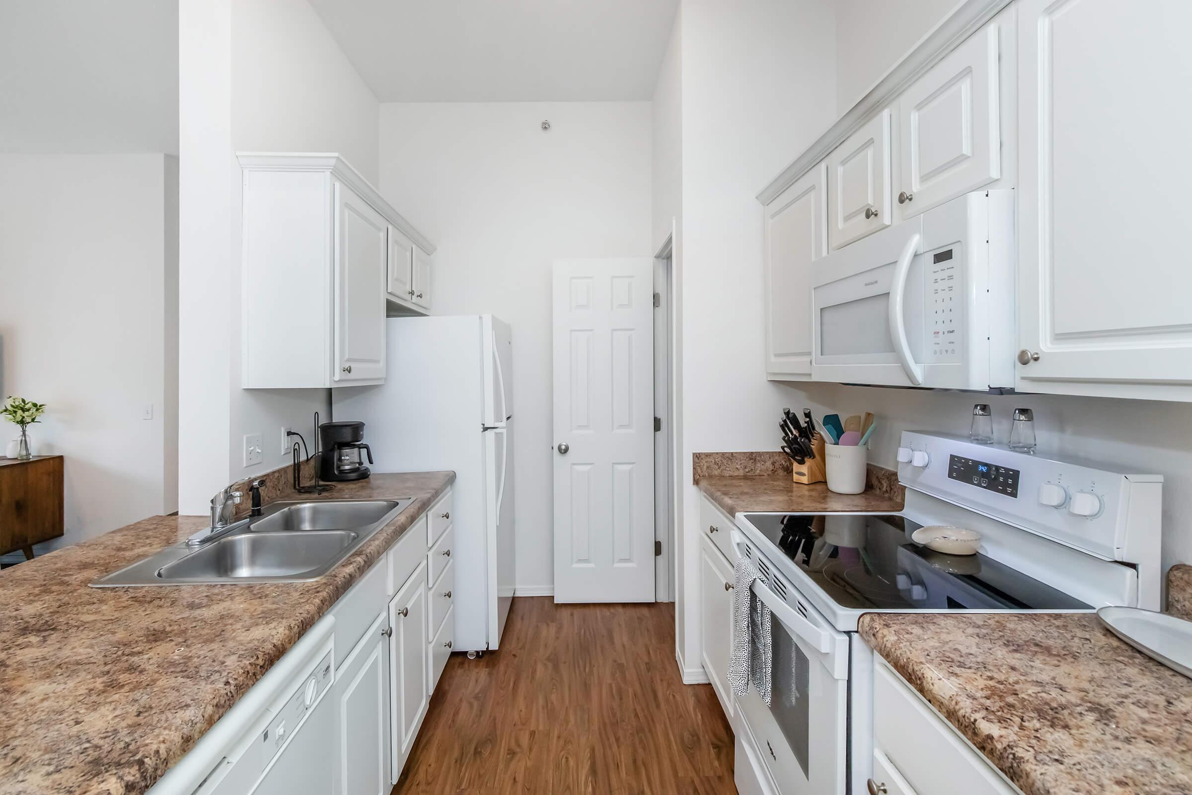 Modern kitchen featuring white cabinetry, a double sink, and granite countertops. Equipped with a stove, microwave, and refrigerator. The space is well-lit with neutral-colored walls and wood-like flooring. A door leads to another room, and kitchen utensils are neatly arranged on the counter.