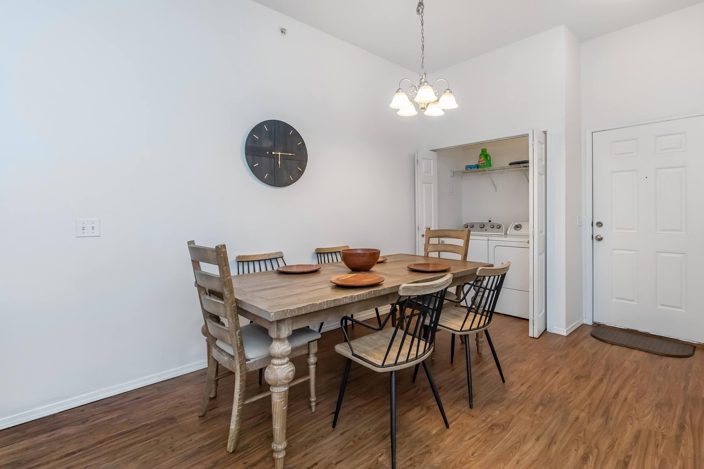 A modern dining area featuring a large wooden table surrounded by six chairs. The room has light-colored walls and a clock on the wall. A doorway leads to a laundry area, and there's a front door visible in the background. The floor is made of wood, creating a warm atmosphere.