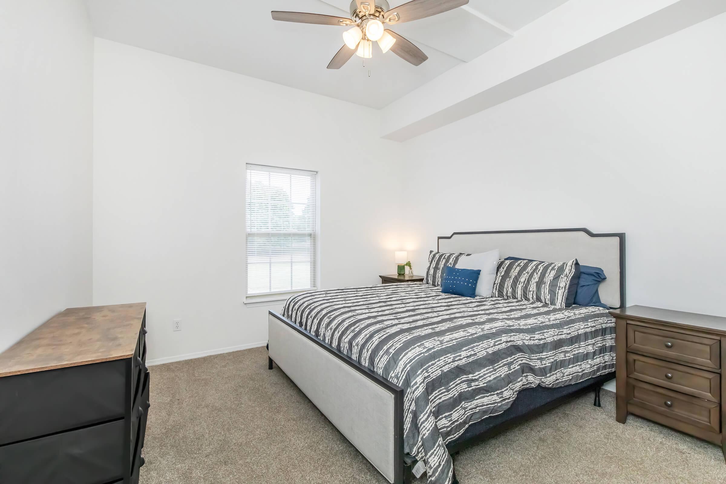 A spacious bedroom featuring a large bed with a striped comforter, two accent pillows, a lamp on a nightstand, and a dresser. The room has light-colored walls, a ceiling fan, and a window that lets in natural light, enhancing the clean and inviting atmosphere.