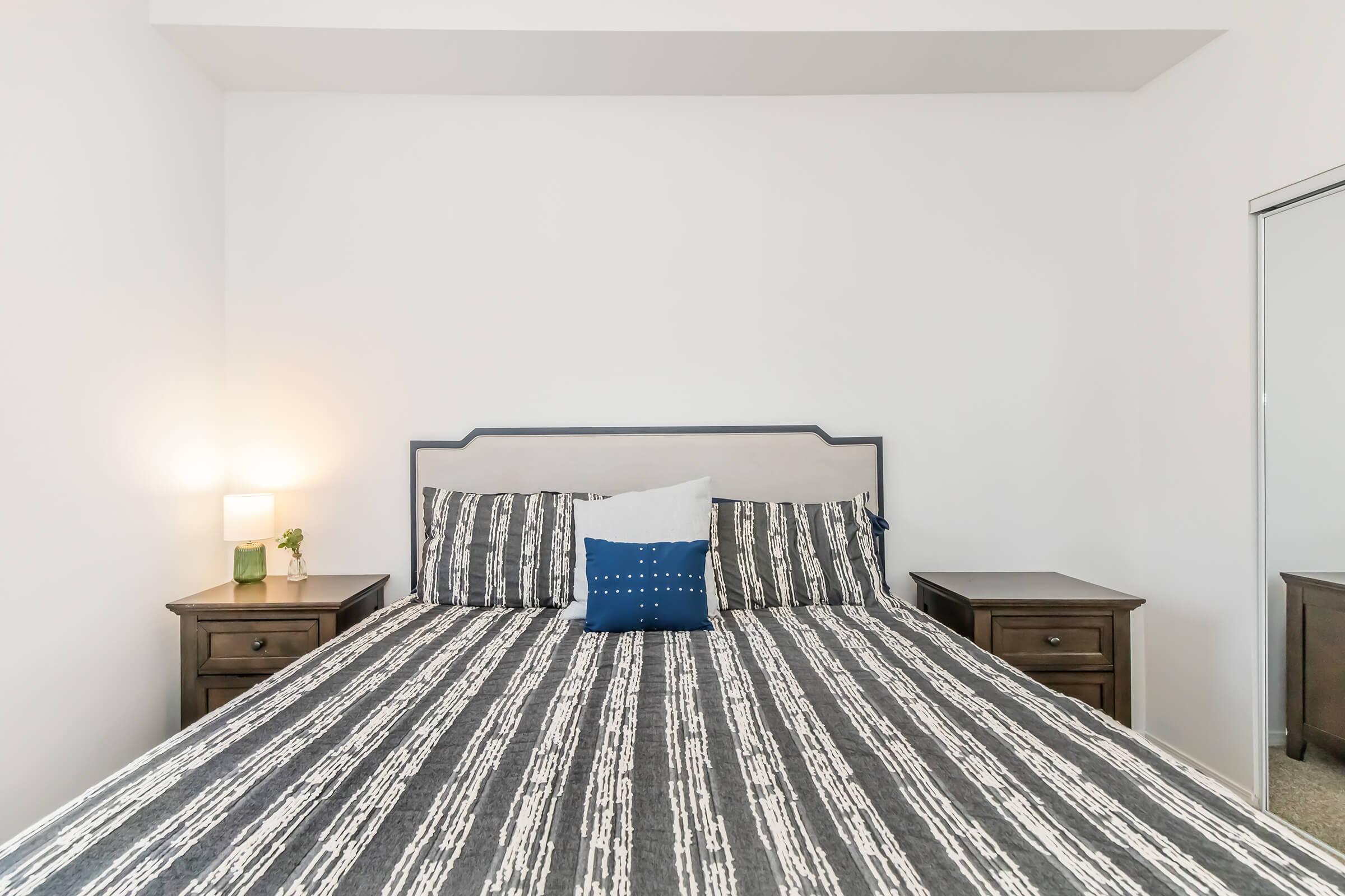 A modern bedroom featuring a neatly made bed with a striped gray and white bedding set. There are two bedside tables, each with a lamp. A decorative blue pillow is placed on the bed. A mirrored closet can be seen on the right side, and the walls are painted white, creating a clean and simple aesthetic.