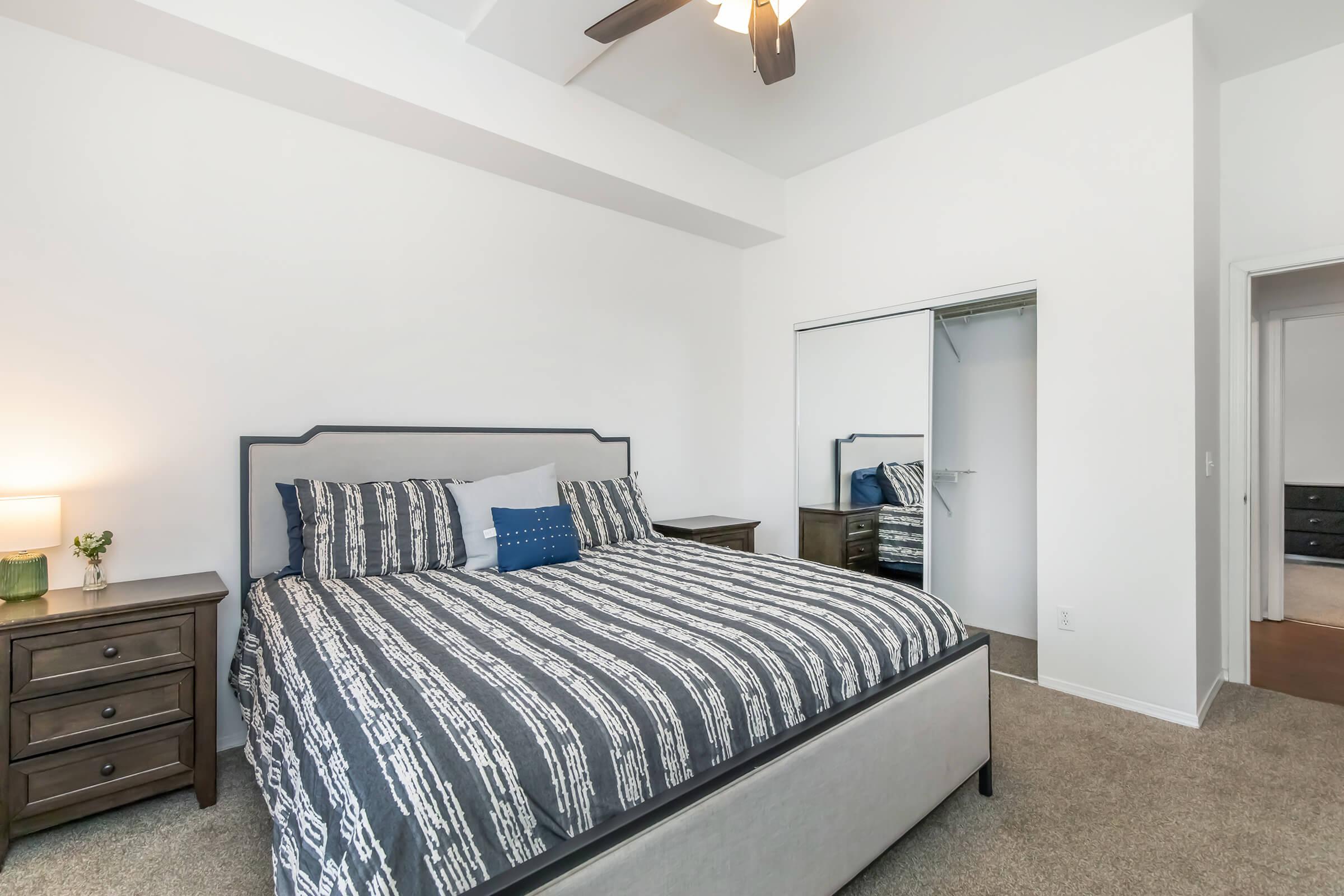 A well-decorated bedroom featuring a queen-sized bed with a patterned comforter, two bedside tables with lamps, and a ceiling fan. There's a closet with mirrored doors and soft carpet flooring. The walls are painted white, giving the room a bright and open feel.