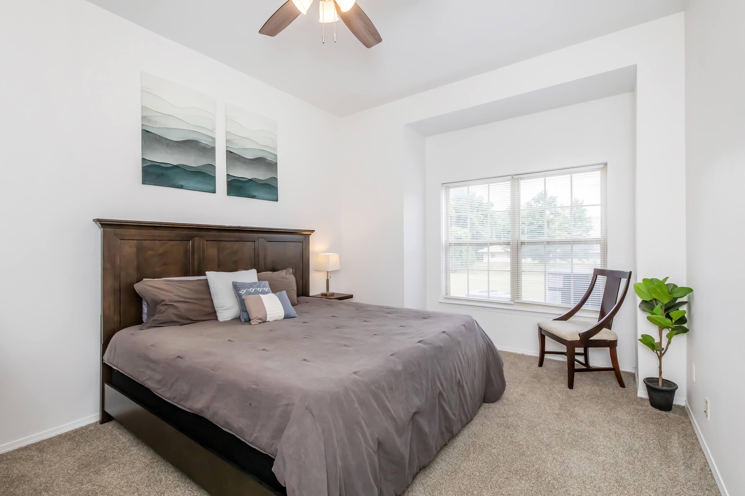 A well-decorated bedroom featuring a large bed with gray bedding, two abstract art pieces on the wall, a wooden headboard, a lamp on a nightstand, a window with white blinds, and a plant beside a wooden chair. The carpeted floor adds warmth to the space.